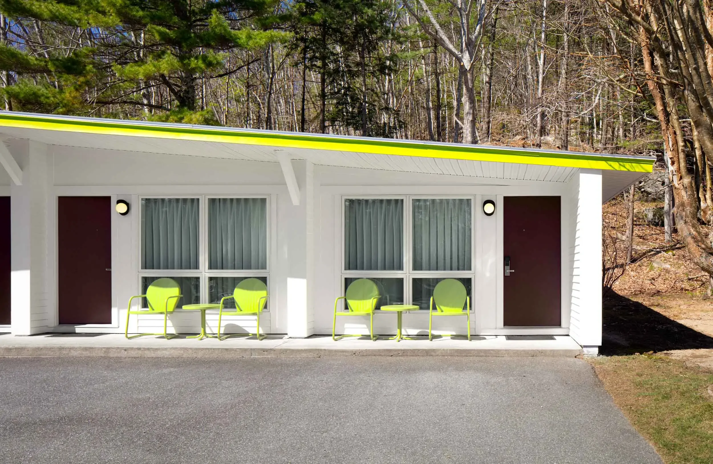 Front view of a modern motel room with a patio area, four lime-green chairs, a small white table, large windows with white curtains, two dark brown doors, and a yellow-green striped roof edge, with a wooded hillside background.