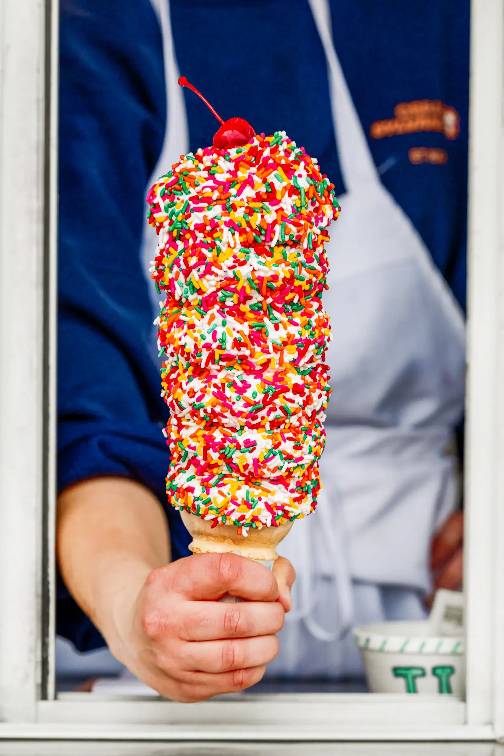 Person holding a colorful ice cream cone with rainbow sprinkles and a cherry on top, seen through a window.