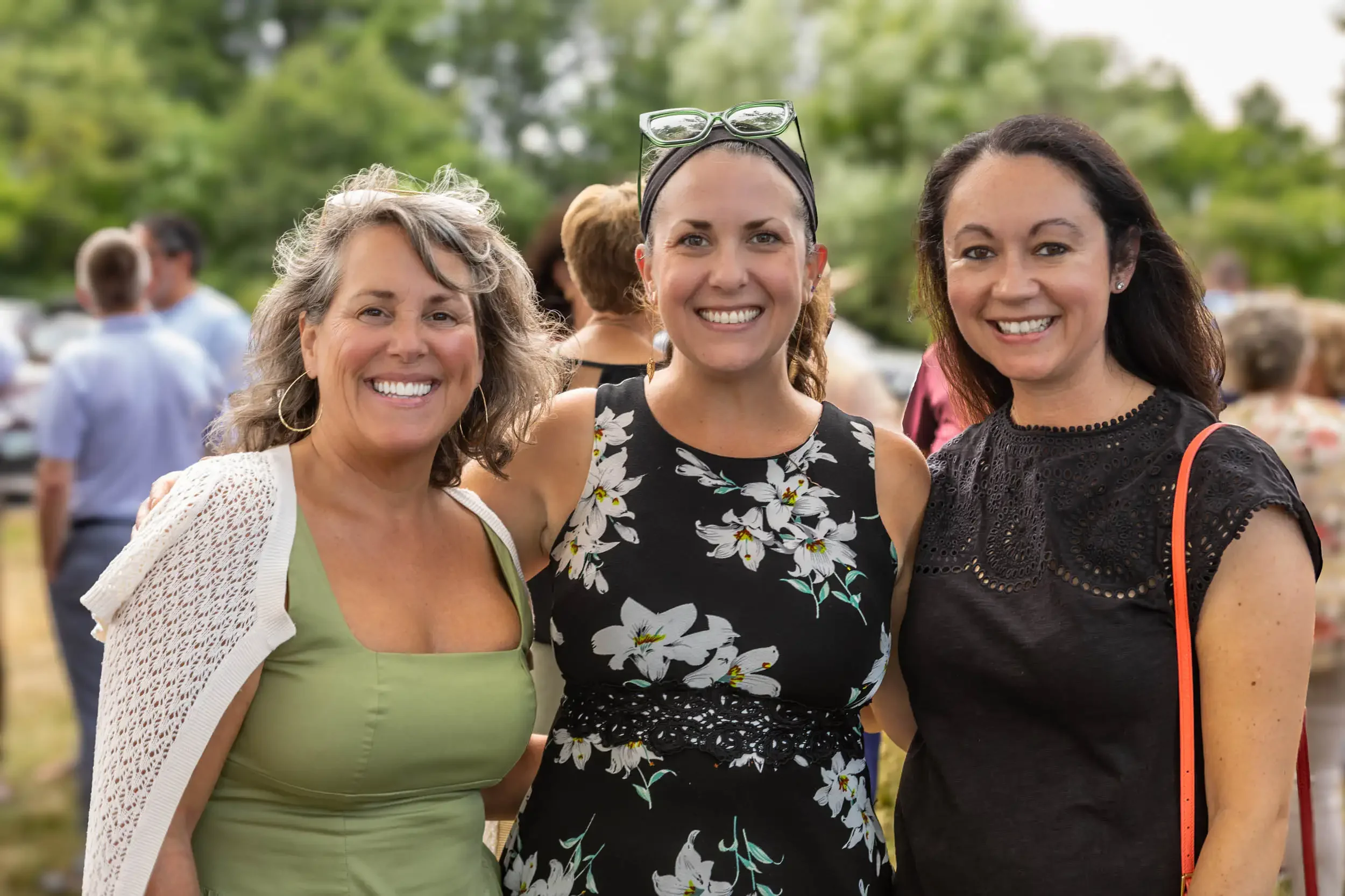 Three women smiling at an outdoor gathering, with greenery and other people in the background.