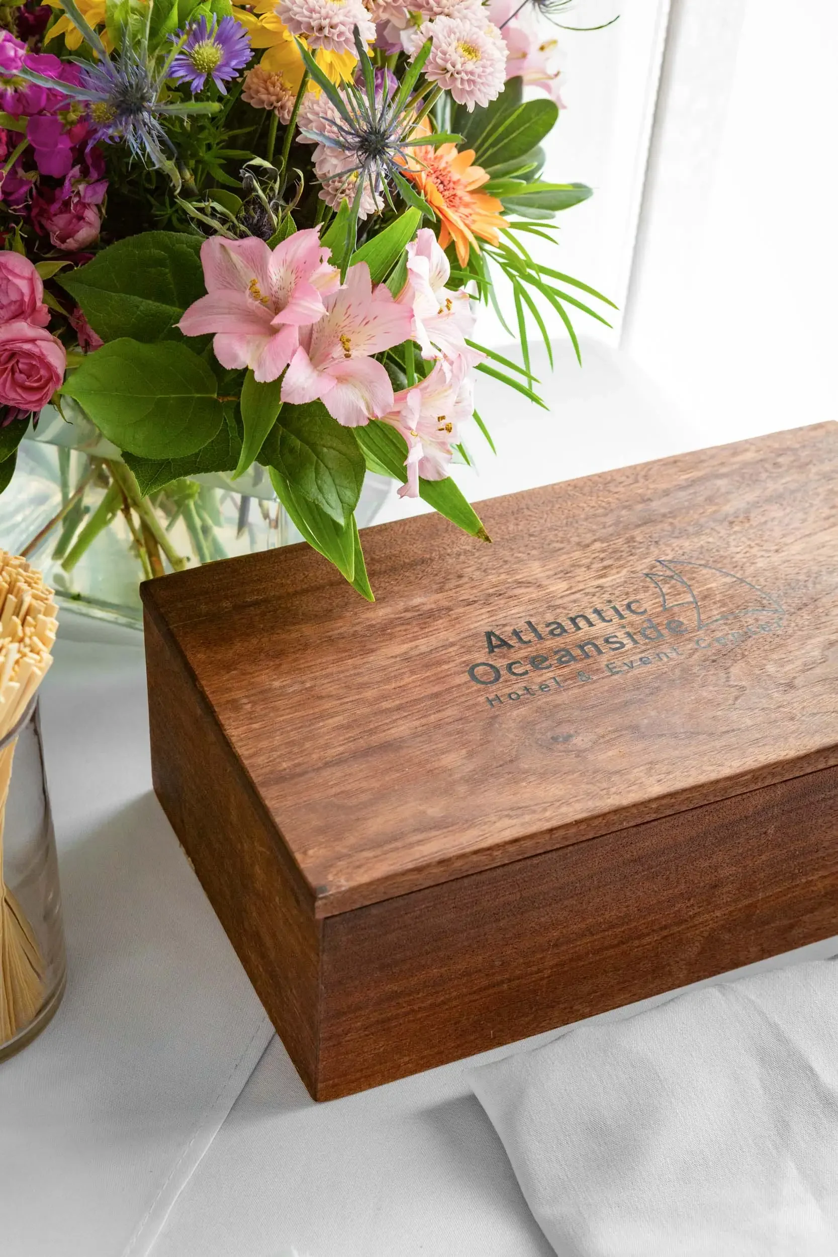 A wooden box with the logo for Atlantic Oceanside Hotel & Event Center on top. Behind it, a bouquet of flowers with pink, orange, yellow, purple, and green leaves is visible.