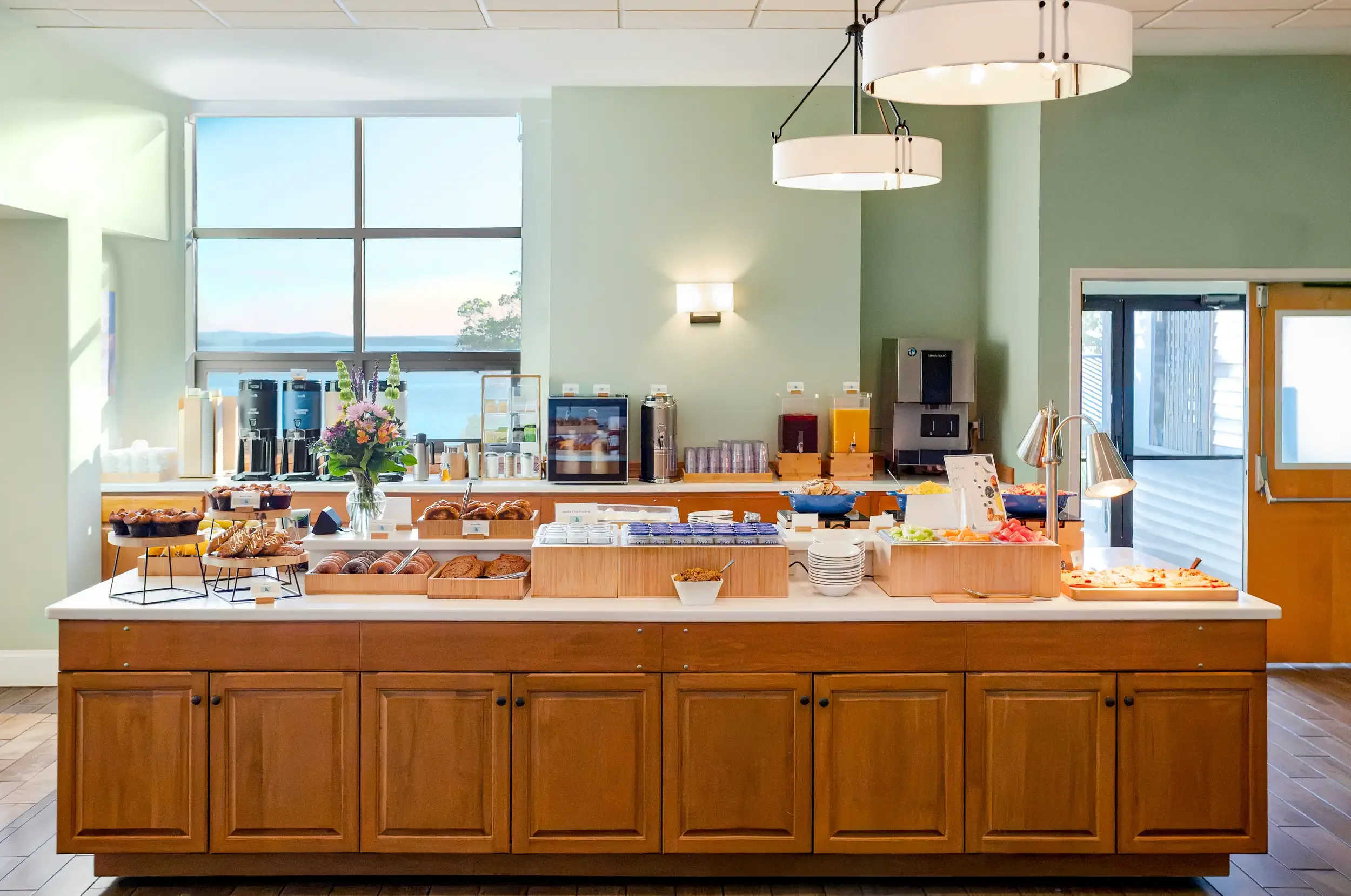 Buffet table with baked goods, fresh fruit, plates, cups, and beverages in a bright dining area with large windows and modern decor.