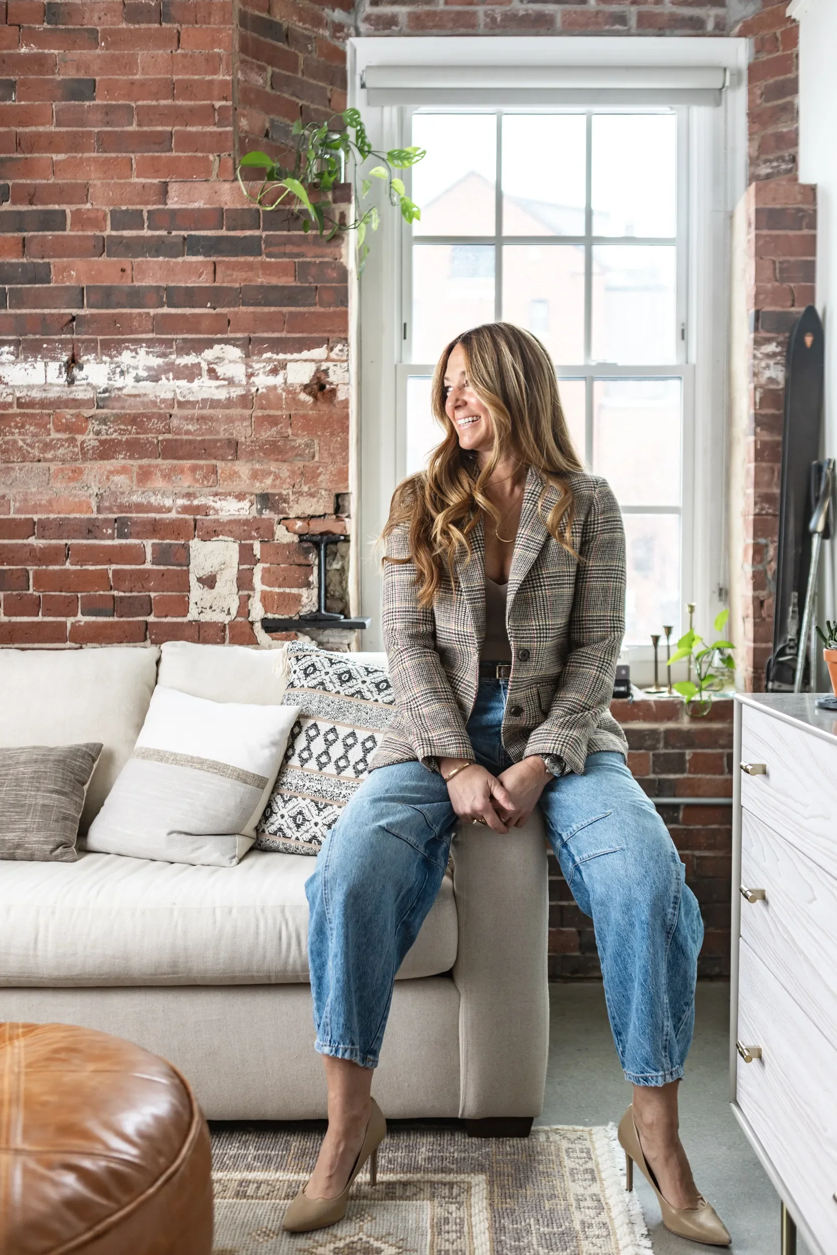 A woman with long, wavy hair wearing a plaid blazer and jeans sitting on a ottoman in a room with exposed brick walls and a large window, smiling and looking to her right. There are pillows on a beige sofa, a leather chair in the foreground, and a wh