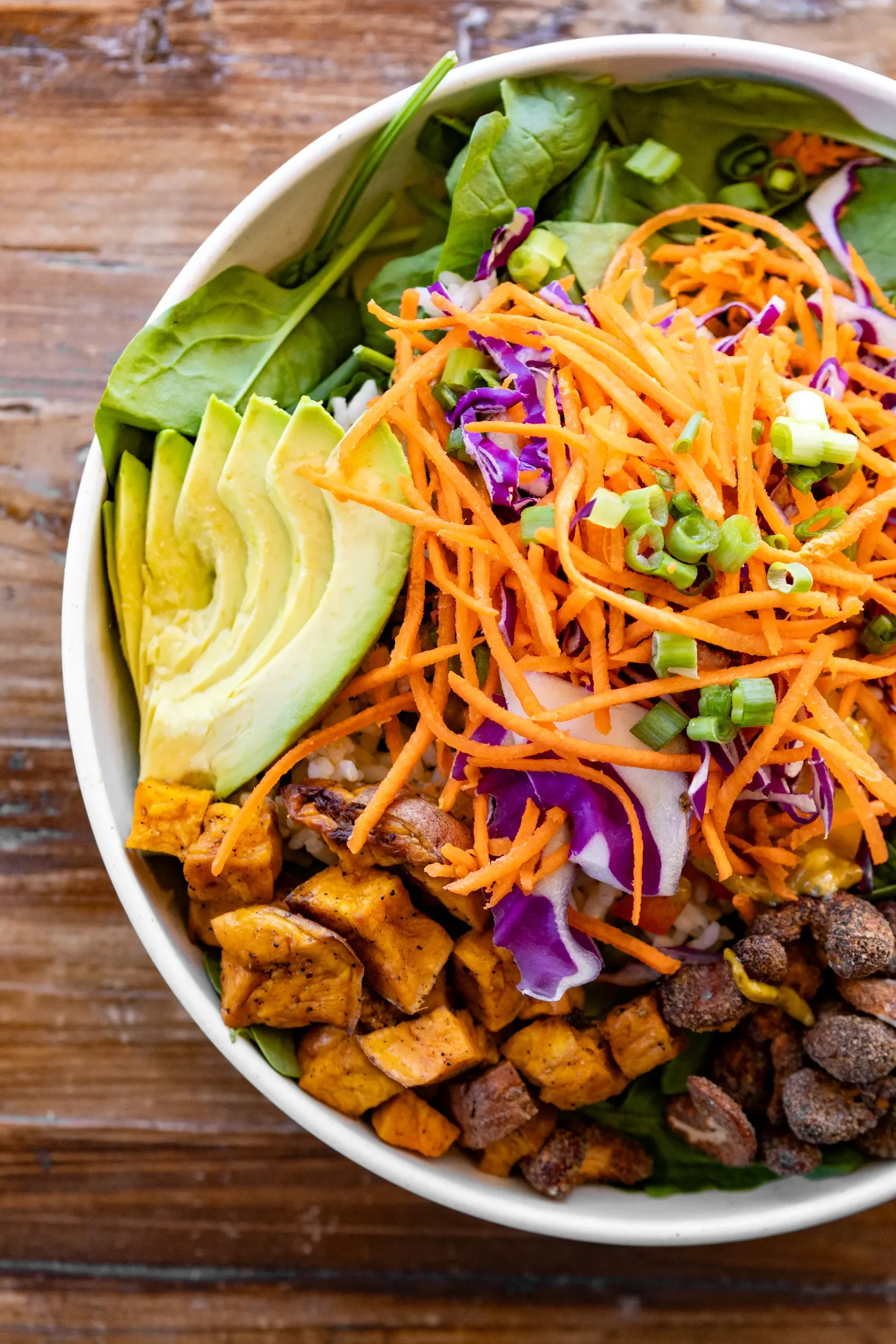 A white bowl of salad containing shredded carrots, purple cabbage, sliced avocado, chopped green onions, mixed greens, grilled chicken, and crumbled bacon on a wooden surface.