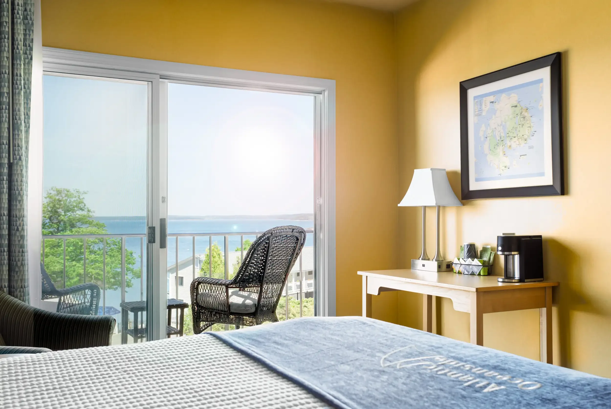 Hotel room with sliding glass door leading to balcony, ocean view, bed in foreground, yellow walls, framed map or artwork, lamp, and coffee station.