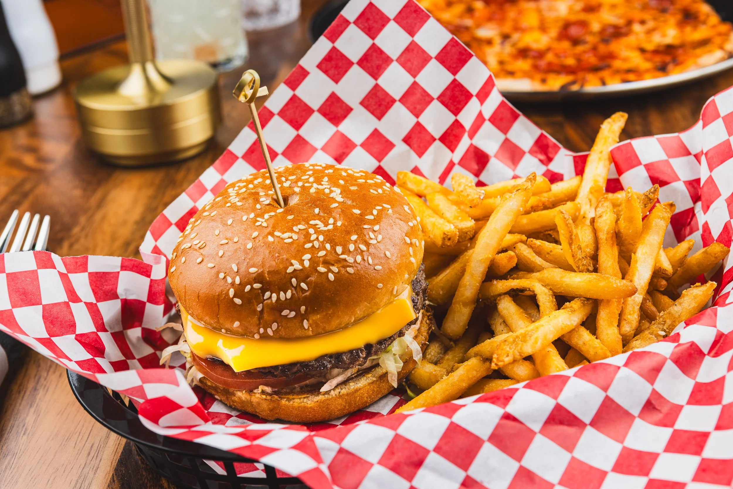 A cheeseburger with lettuce, tomato, and cheese in a sesame seed bun, served with a side of French fries in a black basket lined with red and white checkered paper on a wooden table.