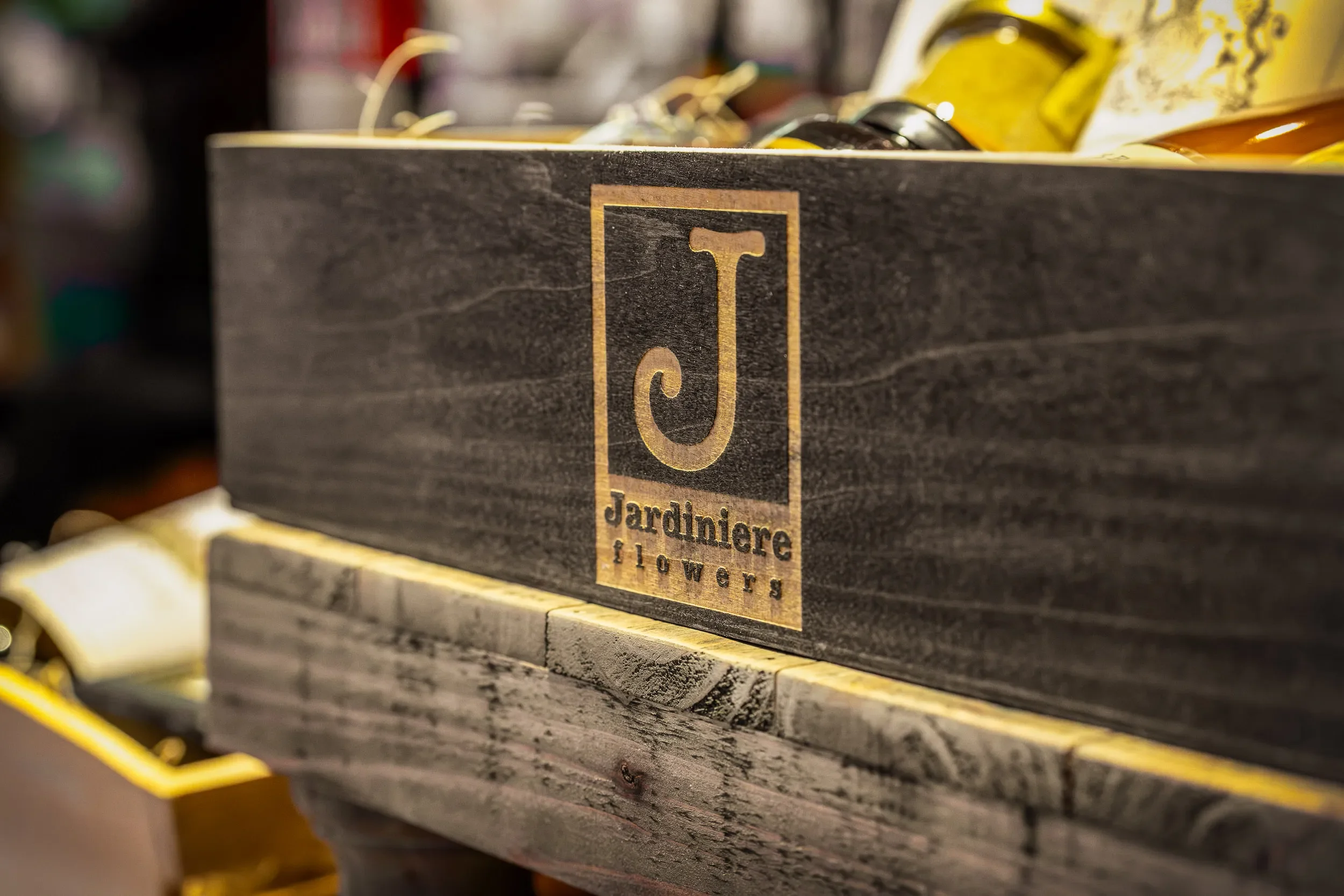 A black wooden box with a gold label featuring a letter 'J' and the words 'Jardiniere flowers' in black text.