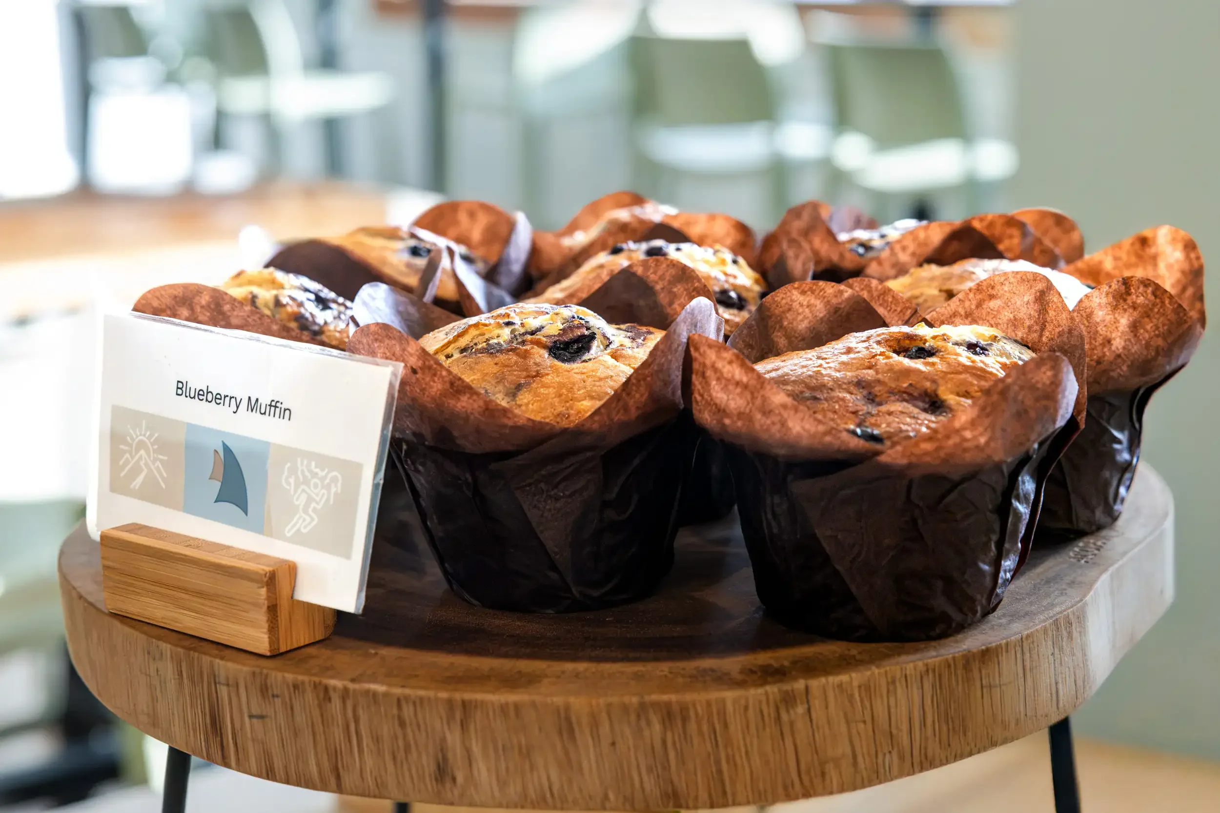 A tray of blueberry muffins on a wooden table, with a small sign card in front labeled 'Blueberry Muffin' and symbols indicating heat and freshness, in a bright, indoor setting.