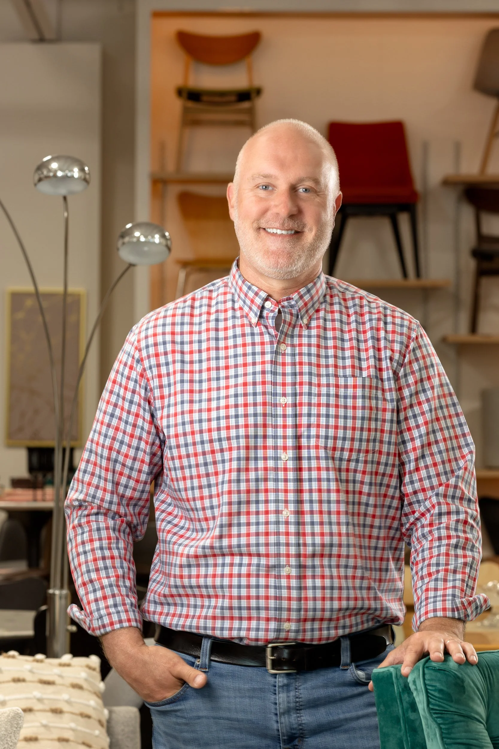 A smiling man with a beard and wearing a red, white, and blue plaid shirt and blue jeans, standing in a furniture store with chairs and lamps in the background.