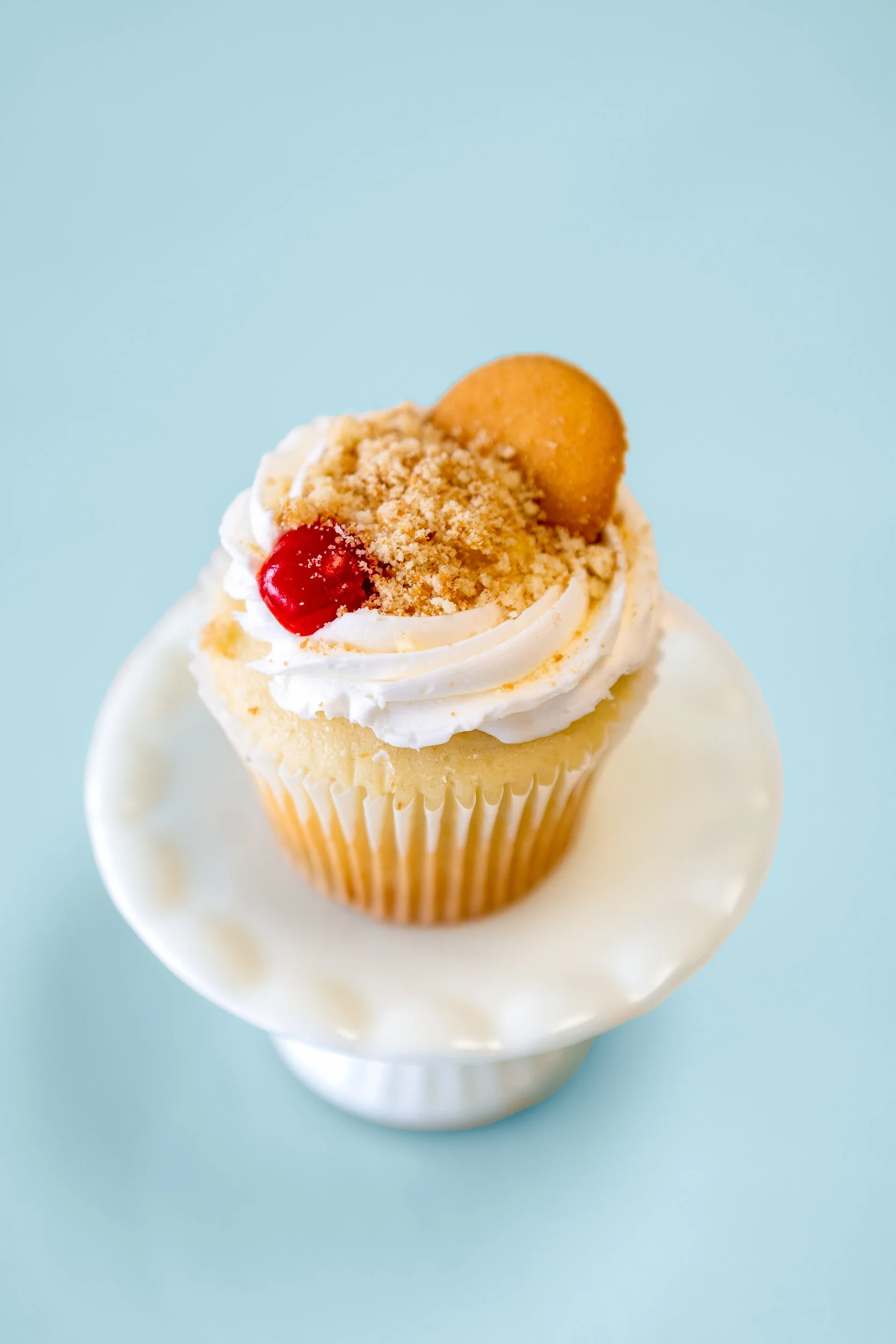 Cupcake topped with whipped cream, cherry, cookie, and crumbled topping on a small white stand against a light blue background.