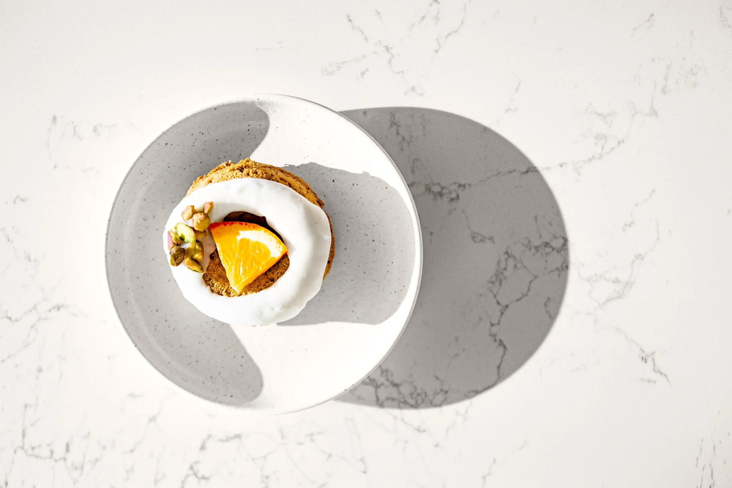 A dessert with a cookie base topped with a swirl of white cream, an orange wedge, chopped pistachios, and a layer of chocolate, served on a white speckled plate on a white marble surface.