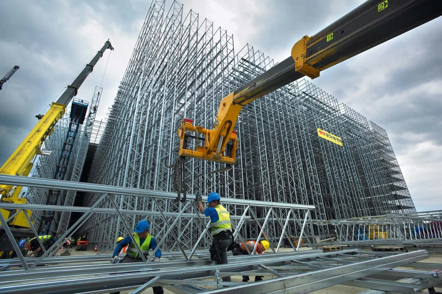 Construction workers in blue and yellow helmets assembling steel framework for a large building, with cranes overhead under cloudy skies.
