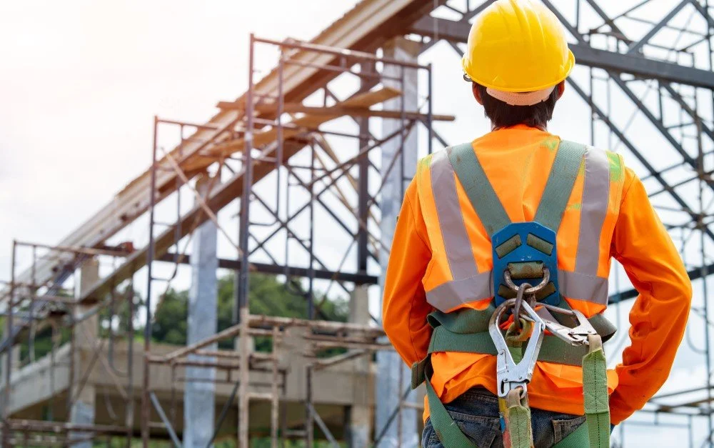 Construction worker wearing a yellow hard hat and orange safety vest observing a building site with scaffolding and structural framework.