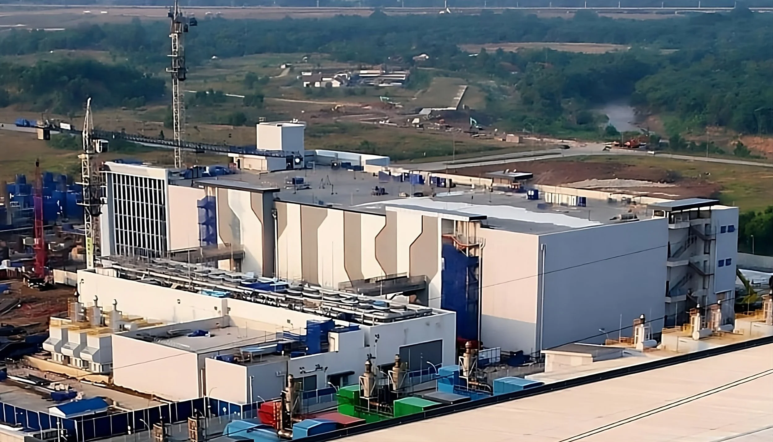 An industrial building with equipment and ventilation systems on the roof, surrounded by construction area, with a rural landscape in the background.