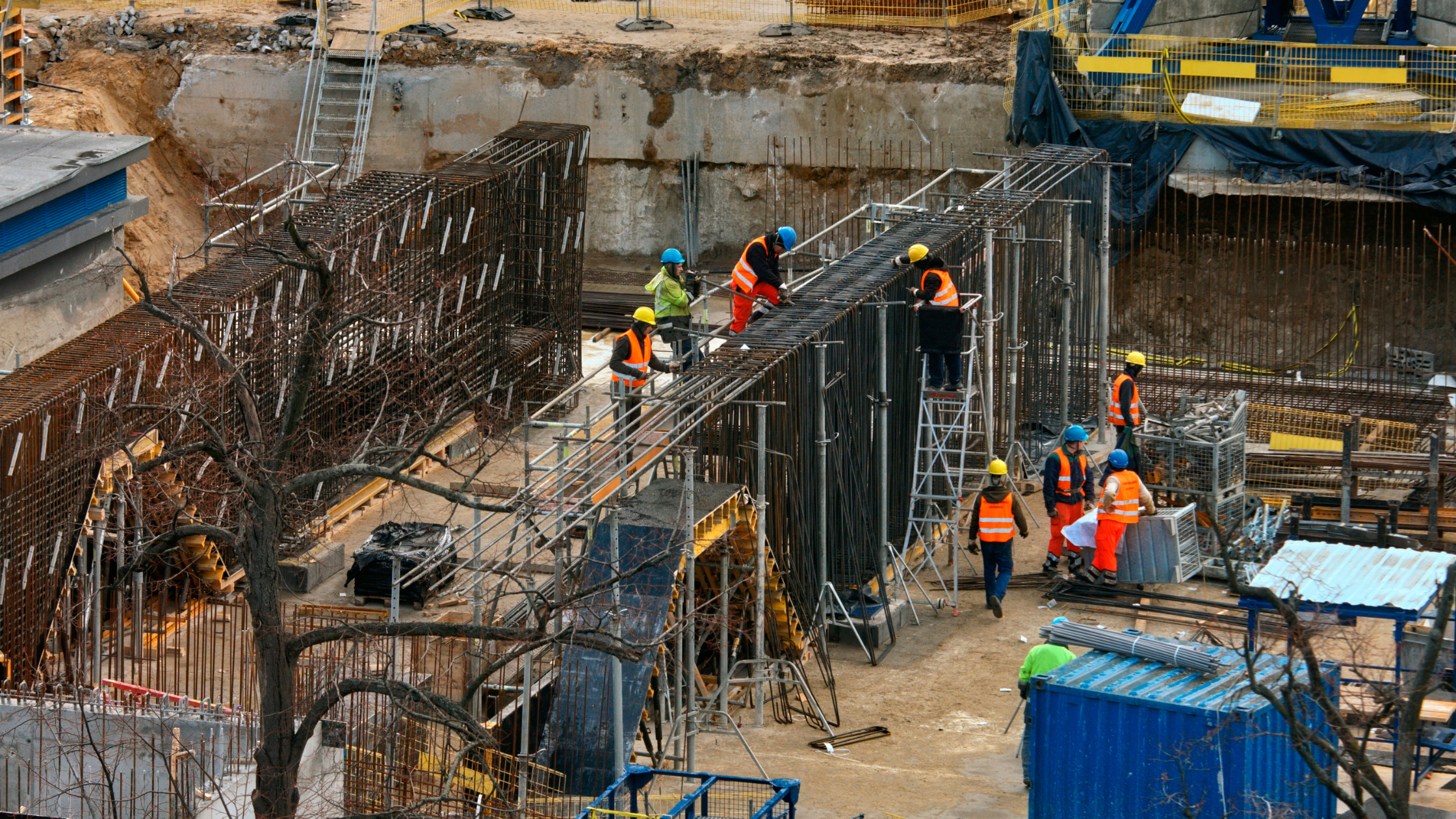 Construction workers wearing safety vests and helmets working on steel reinforcement for a building foundation at a construction site.