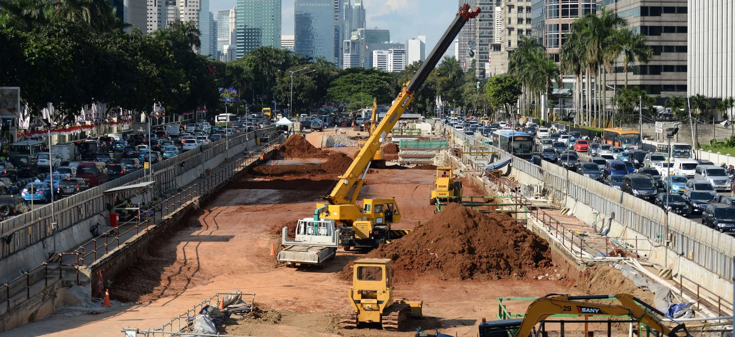 Construction site on a city street with heavy machinery including a crane and excavators, adjacent to a traffic jam with multiple lanes of cars and tall buildings in the background.