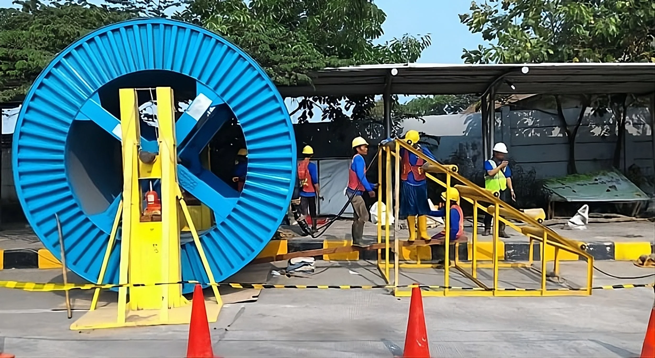 Workers in safety helmets and vests operating overground utility equipment, including a large blue and yellow cable reel, at an outdoor construction or maintenance site with safety cones and barriers.