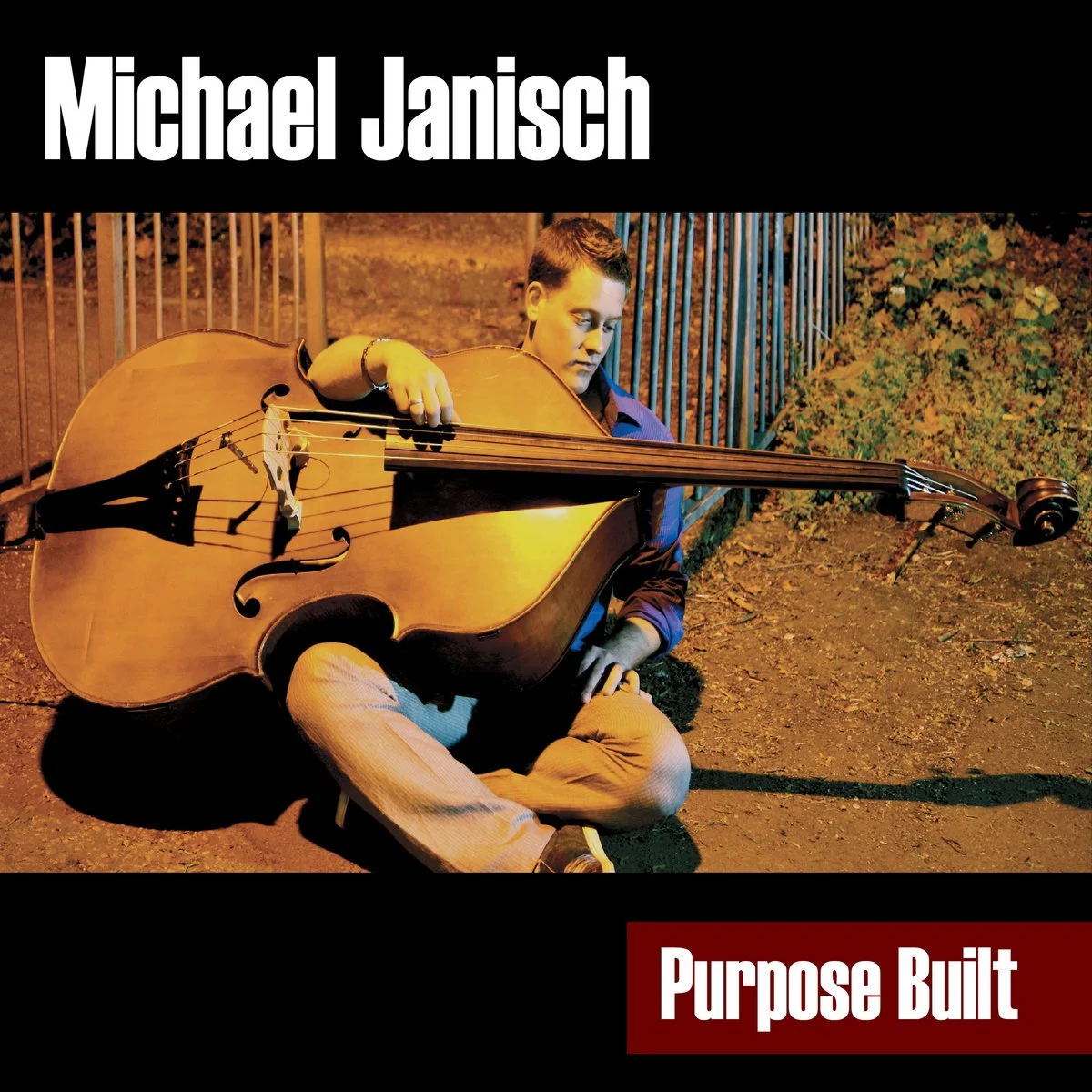 An album cover featuring a young man sitting on the ground outdoors at night, playing a large wooden double bass, with a fence and foliage in the background, and the text 'Michael Janisch' at the top and 'Purpose Built' at the bottom.