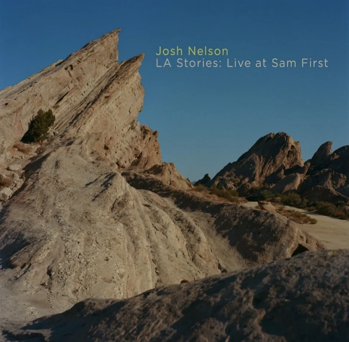Arid desert landscape with large rock formations and mountains under a clear blue sky, with text overlay about Josh Nelson and his documentary 'LA Stories: Live at Sam First'.