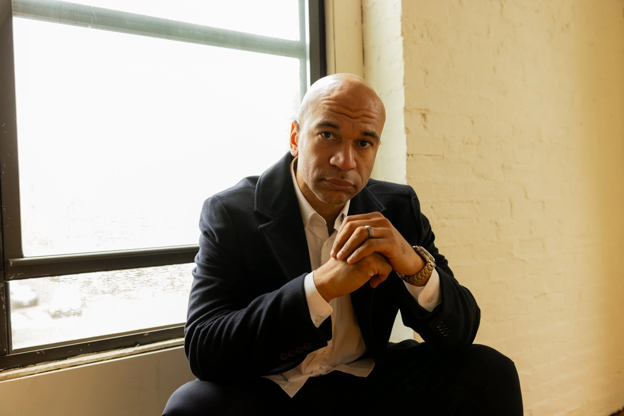 A man with a shaved head wearing a dark blazer and white shirt sitting near a window with a yellow wall behind him, looking at the camera with hands clasped.