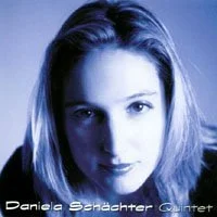 A close-up portrait of a woman with fair skin and straight hair, looking into the camera, with a dark background.