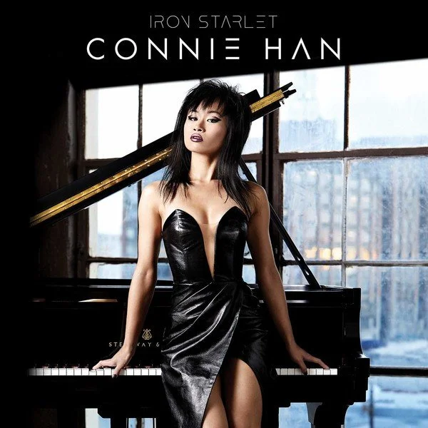 Connie Han sitting at a piano with a city skyline in the background, wearing a black leather dress and holding a golden stick.
