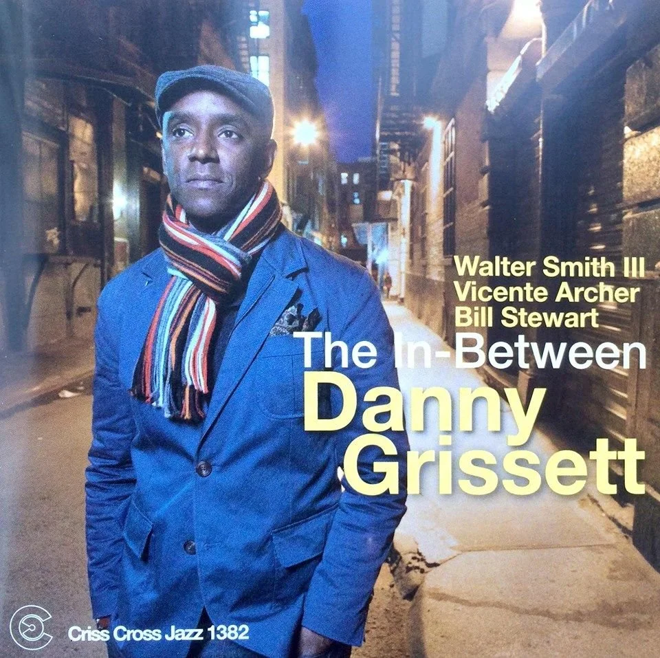 Album cover featuring a man in a blue jacket and flat cap, standing in an urban alley at night, with text highlighting the names Walter Smith III, Vicent Archer, and Bill Stewart, and the album title 'The In-Between Danny Grissett'.