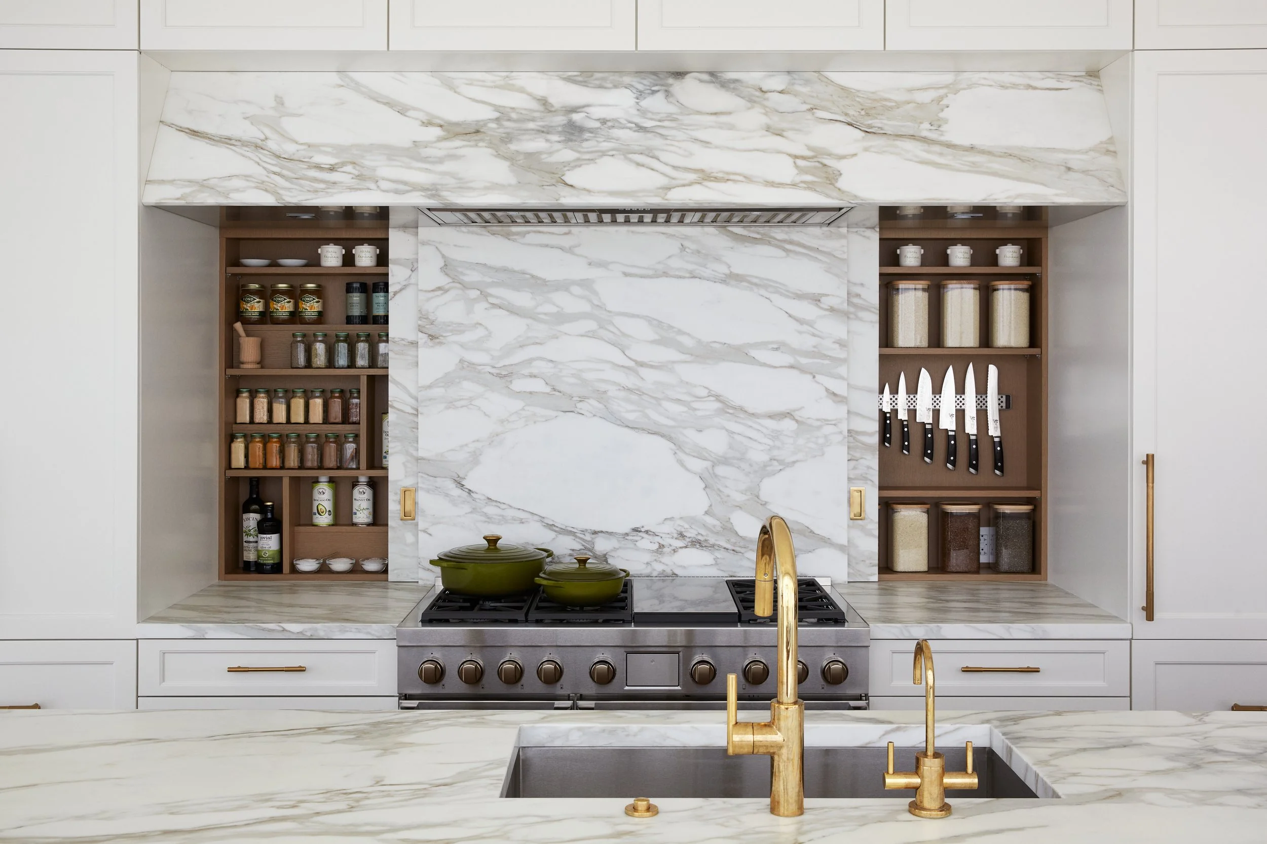 Kitchen with marble backsplash, counters, and island, gold faucet, open shelves with spices, knives, jars, and white dishes, stainless steel stove with green pot and pan.