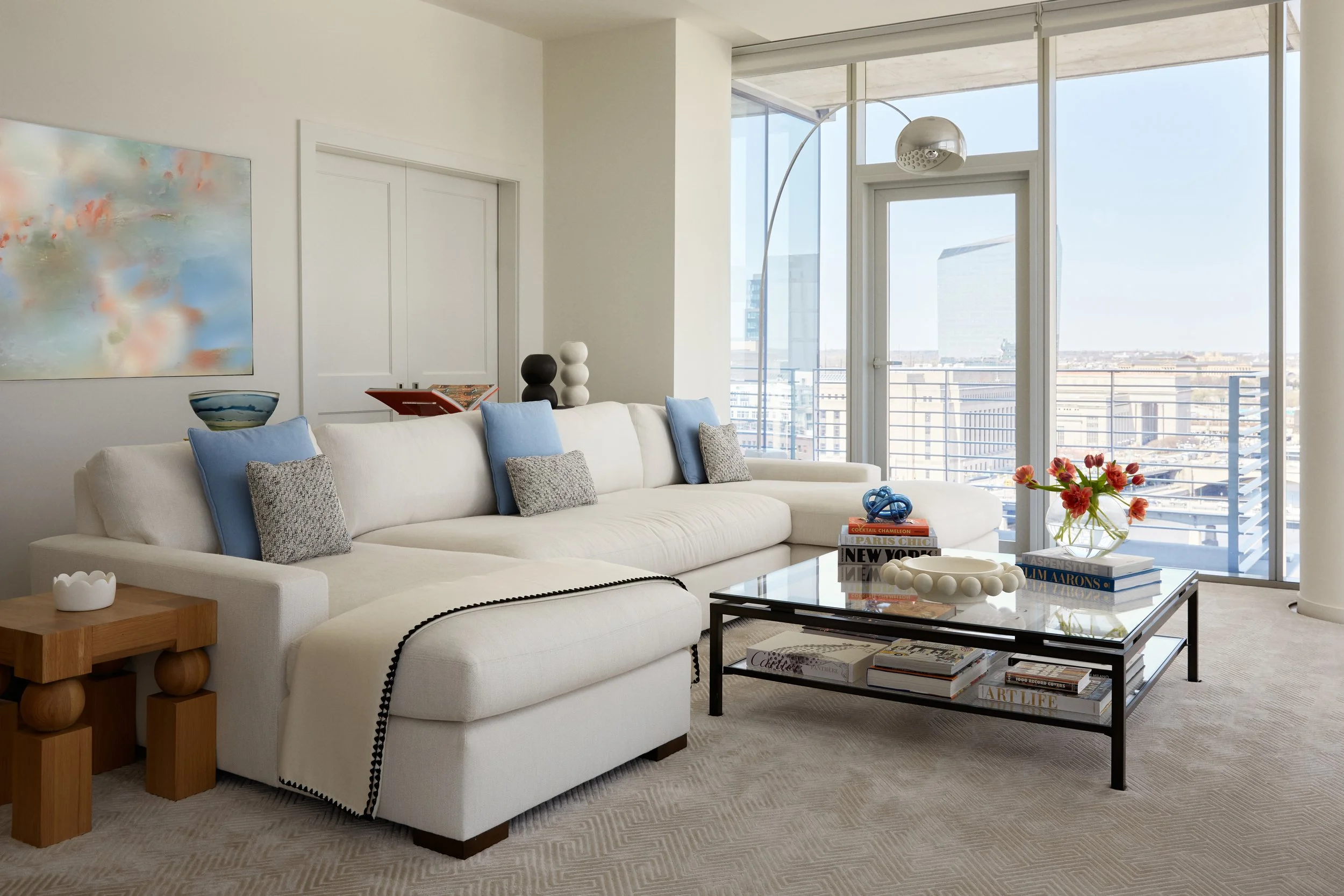 Modern living room with white sectional sofa, colorful pillows, glass coffee table with books and decorative objects, large windows with city view, and minimalist decor.