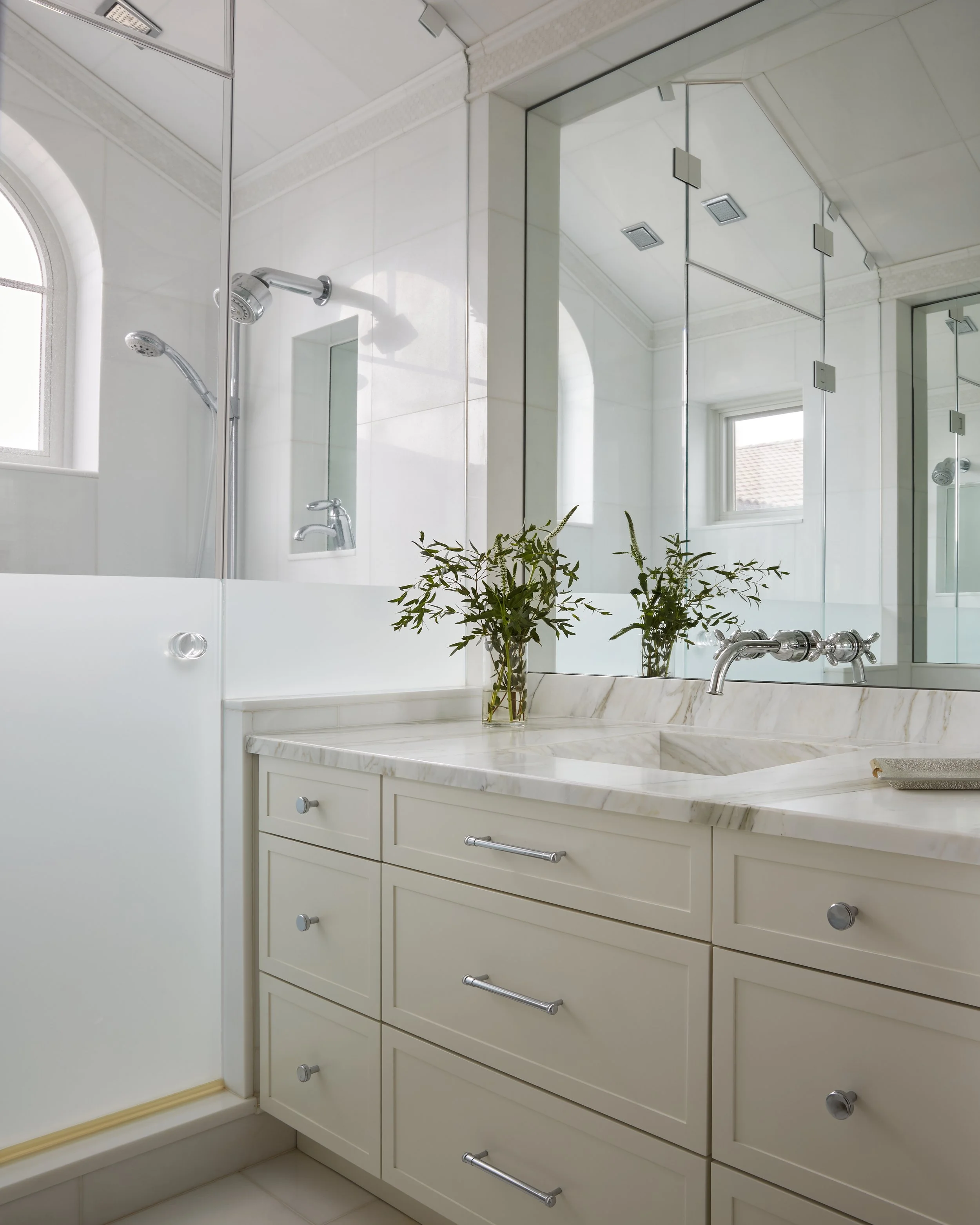 Bathroom with marble countertop, beige cabinets, large mirror, and two small vases of green leafy plants on the counter.