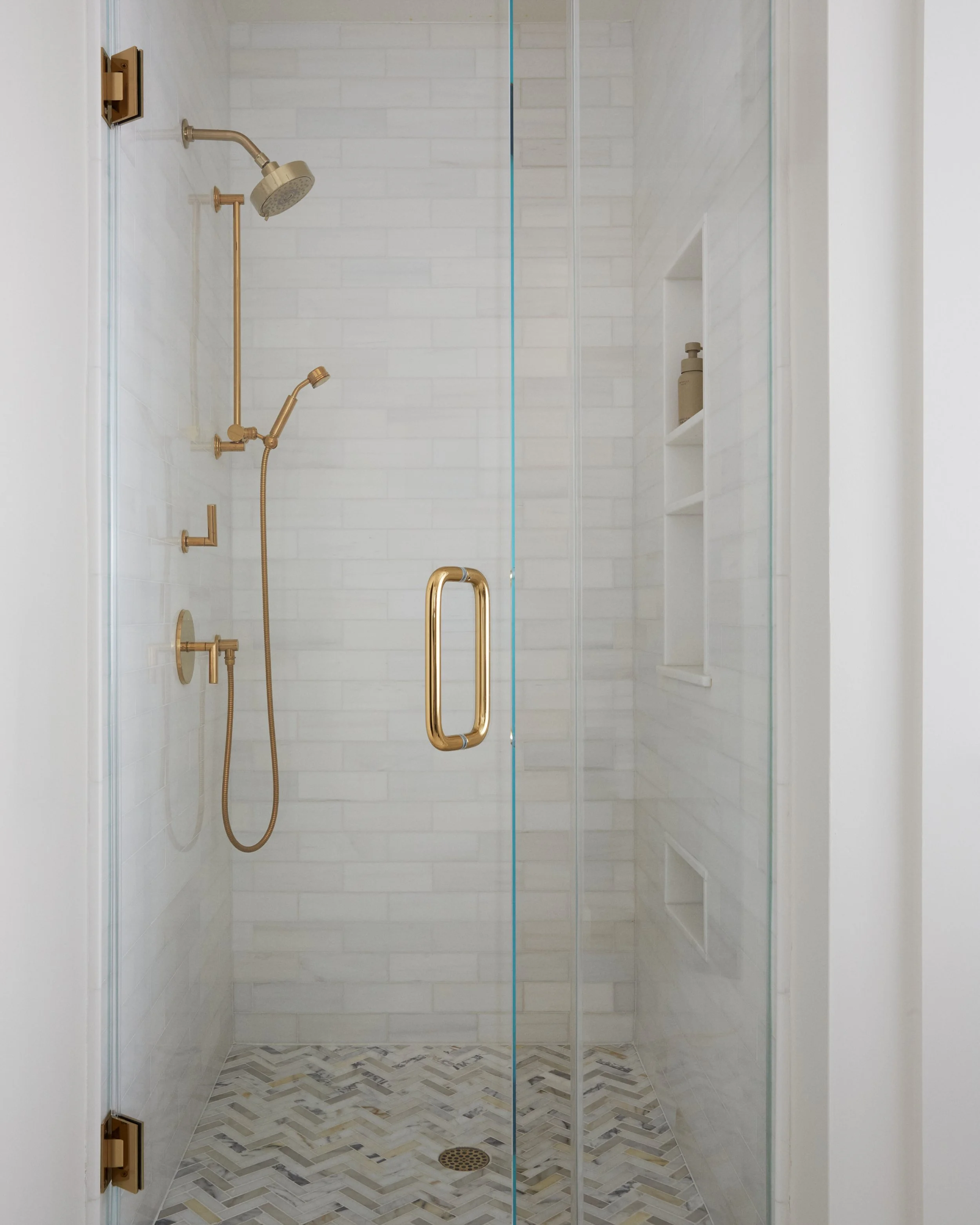 Shower with white subway tiles, gold fixtures, glass door with gold handle, built-in shelves, and chevron-patterned tile floor.