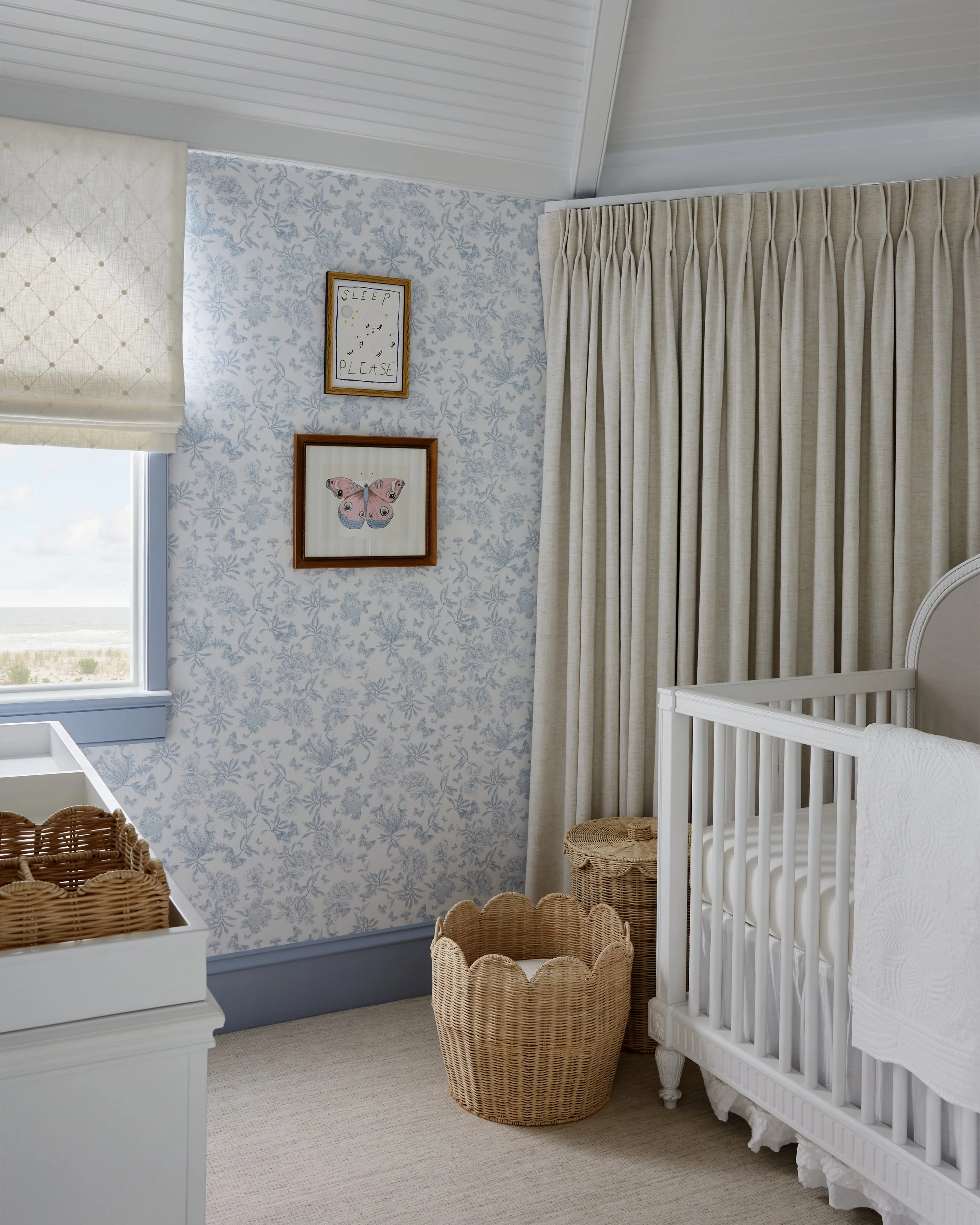 A cozy nursery with white crib, woven baskets, off-white curtains, floral wallpaper, and framed artwork including a butterfly illustration and a sign that says 'Sleep Please'.