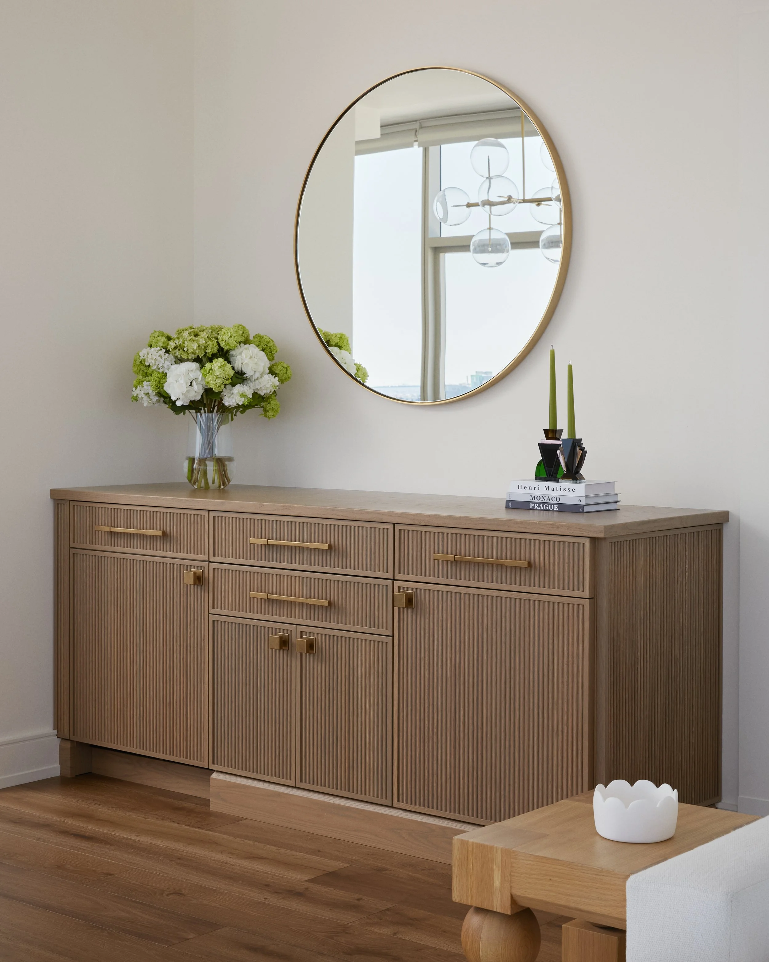 A wooden sideboard with a textured surface and gold handles, decorated with a vase of white and green flowers, a stack of books, and two green candles in black candle holders, with a large round mirror and a chandelier reflected in the background.