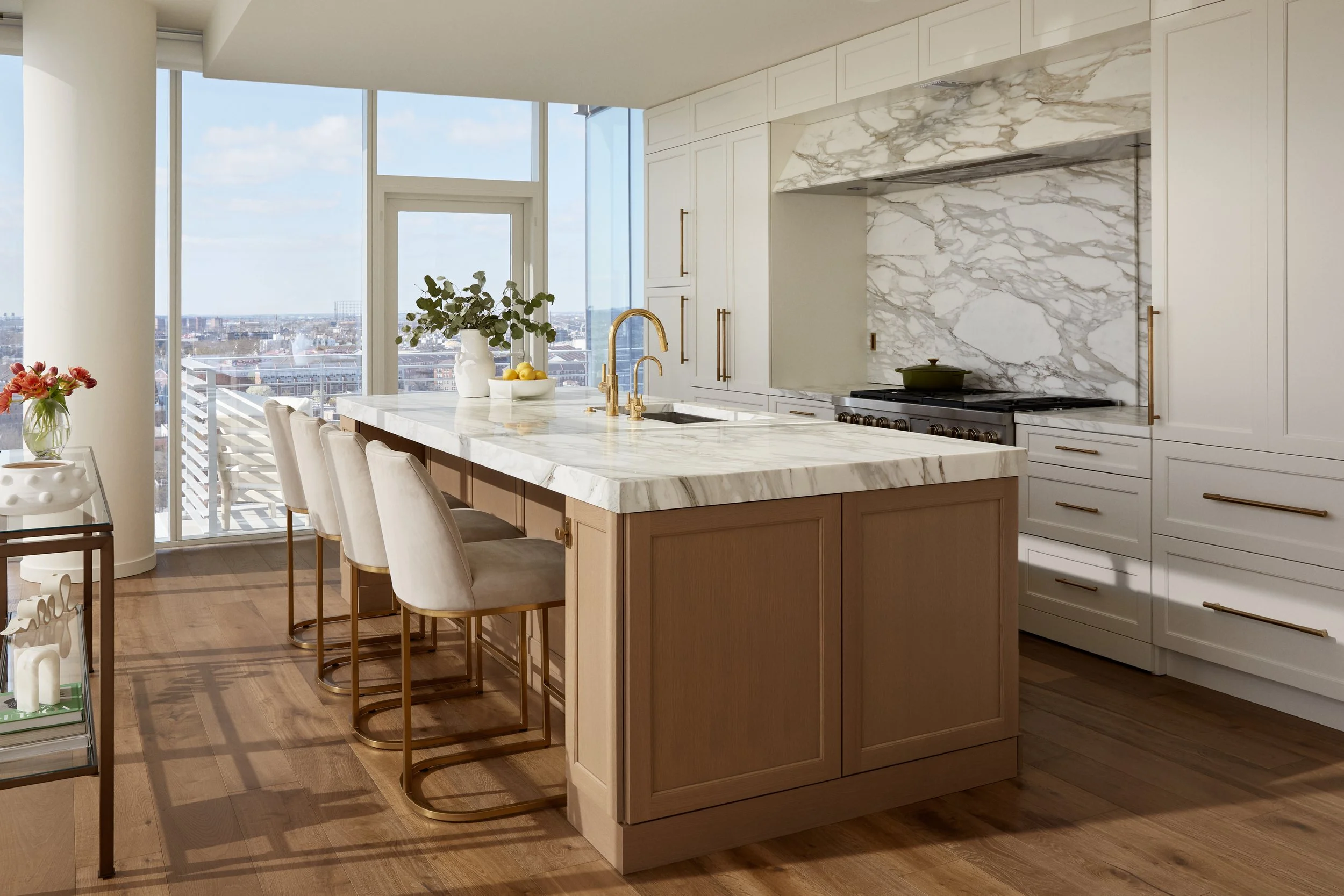 Modern kitchen with large marble island, white cabinets with gold handles, and a marble backsplash. There are four beige upholstered barstools with gold legs, a potted plant, and a view of a cityscape through large windows.
