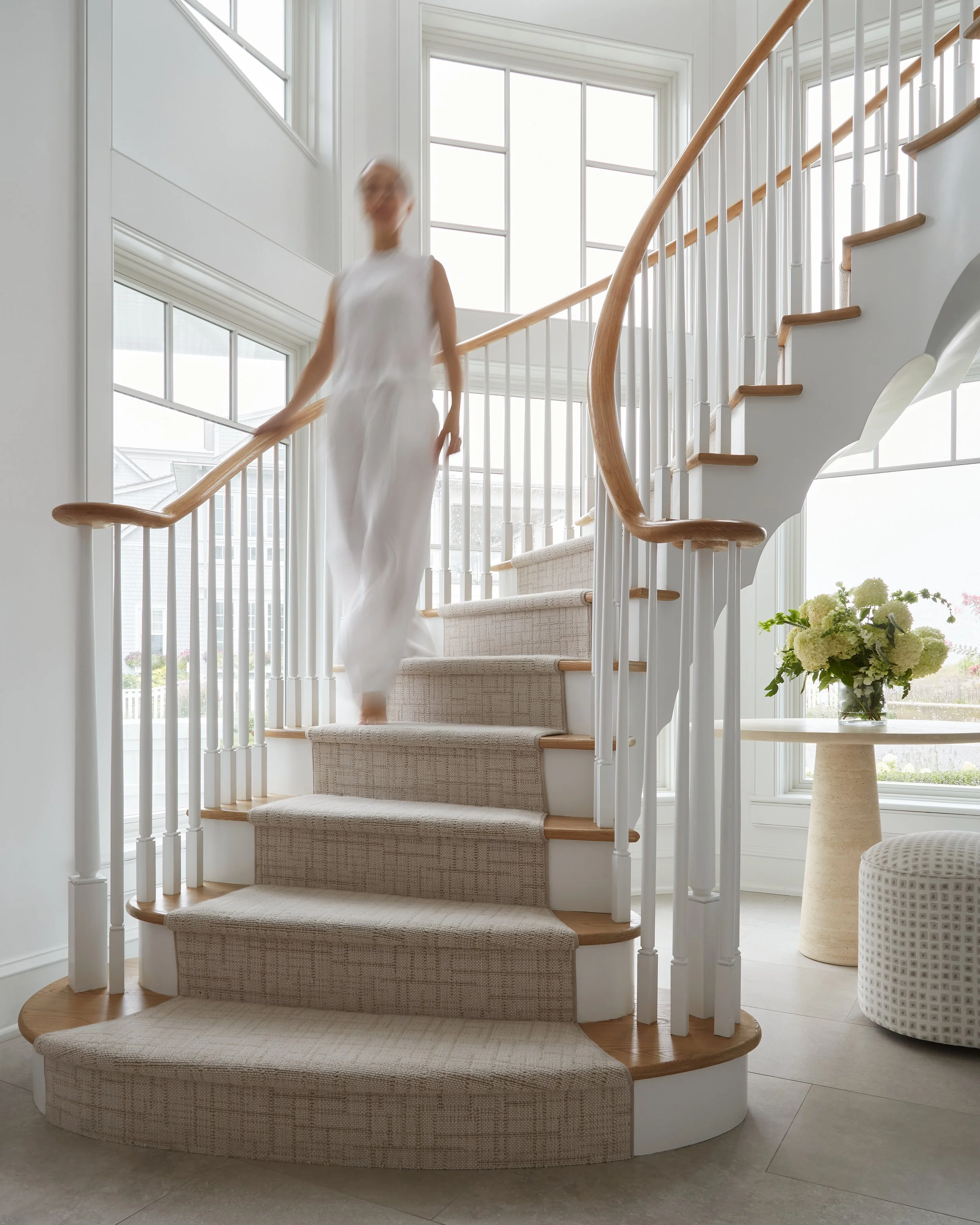 A woman walking down a curved staircase with beige carpet, white railings, and wooden handrails in a bright, modern home with large windows and a flower vase on a side table.