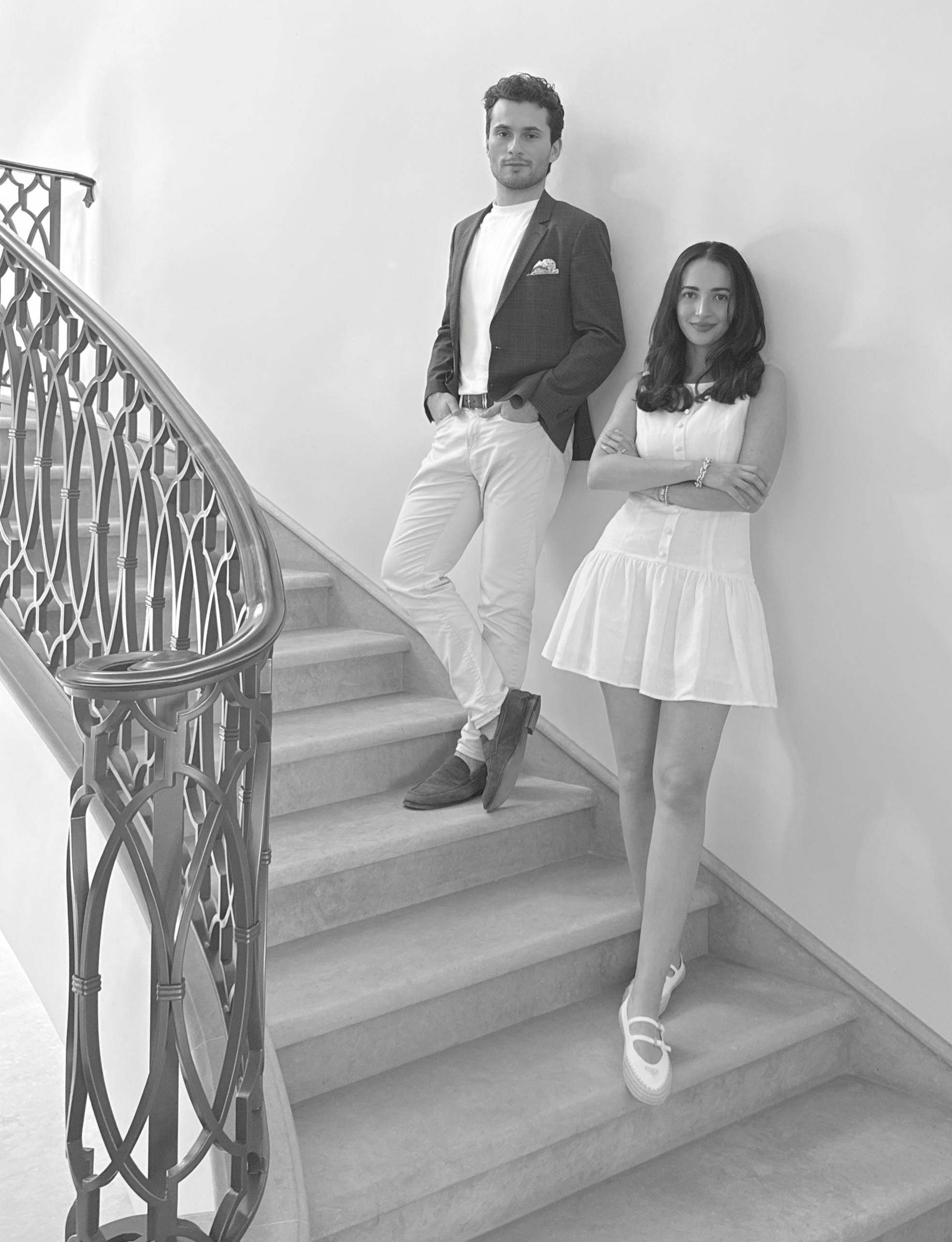 A man and woman standing on a staircase indoors, dressed in semi-formal attire, with the man leaning against the wall and the woman crossing her arms.
