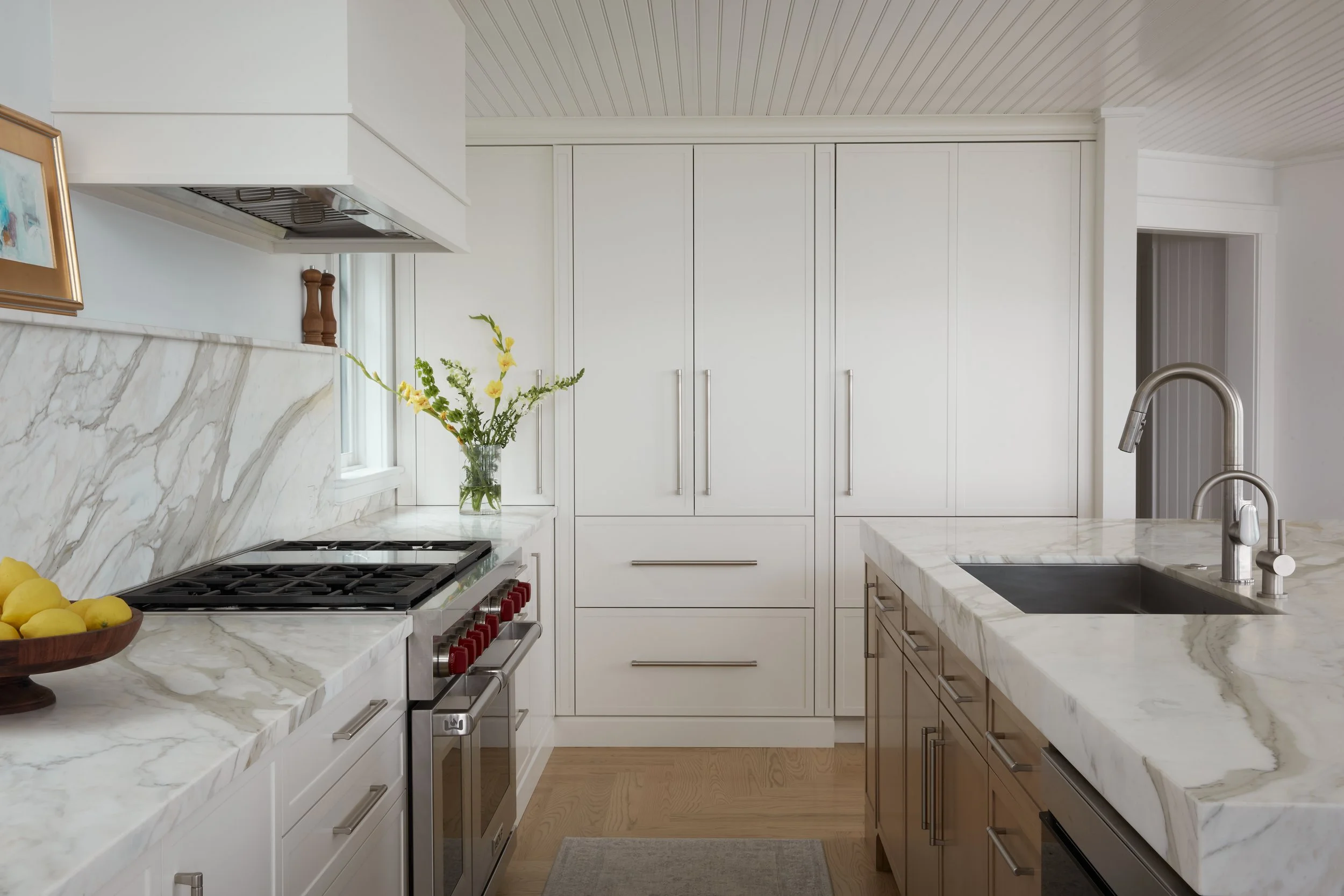 Modern kitchen with white cabinets, marble countertops, a stainless steel oven, a sink with a modern faucet, a vase with yellow flowers, and a bowl of lemons on the counter.