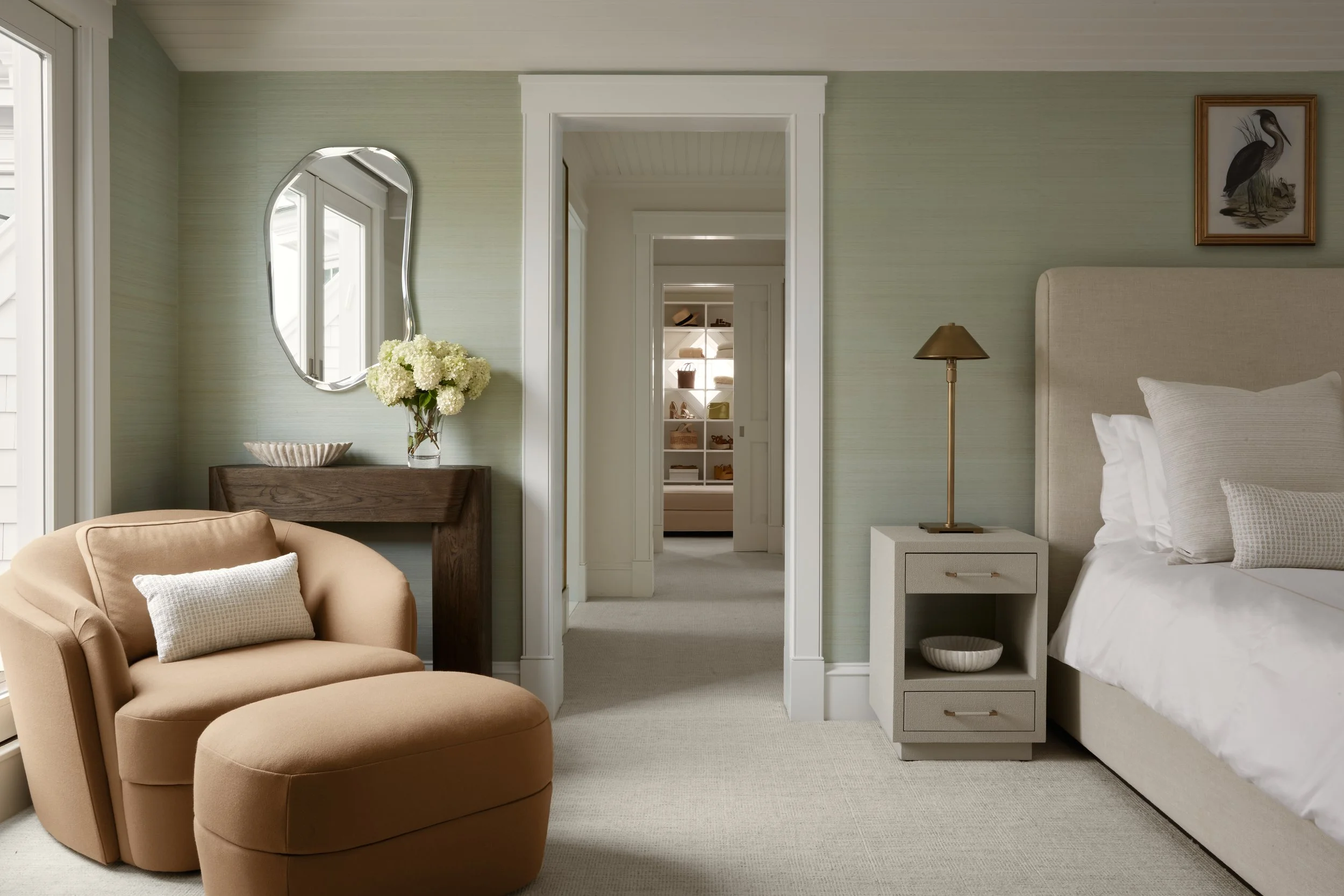 A cozy bedroom with a beige armchair and ottoman, an oval mirror above a dark wood console table with a vase of white flowers, a white nightstand with a gold lamp, a bed with white bedding and pillows, a framed bird picture, and a walk-in closet in the background.