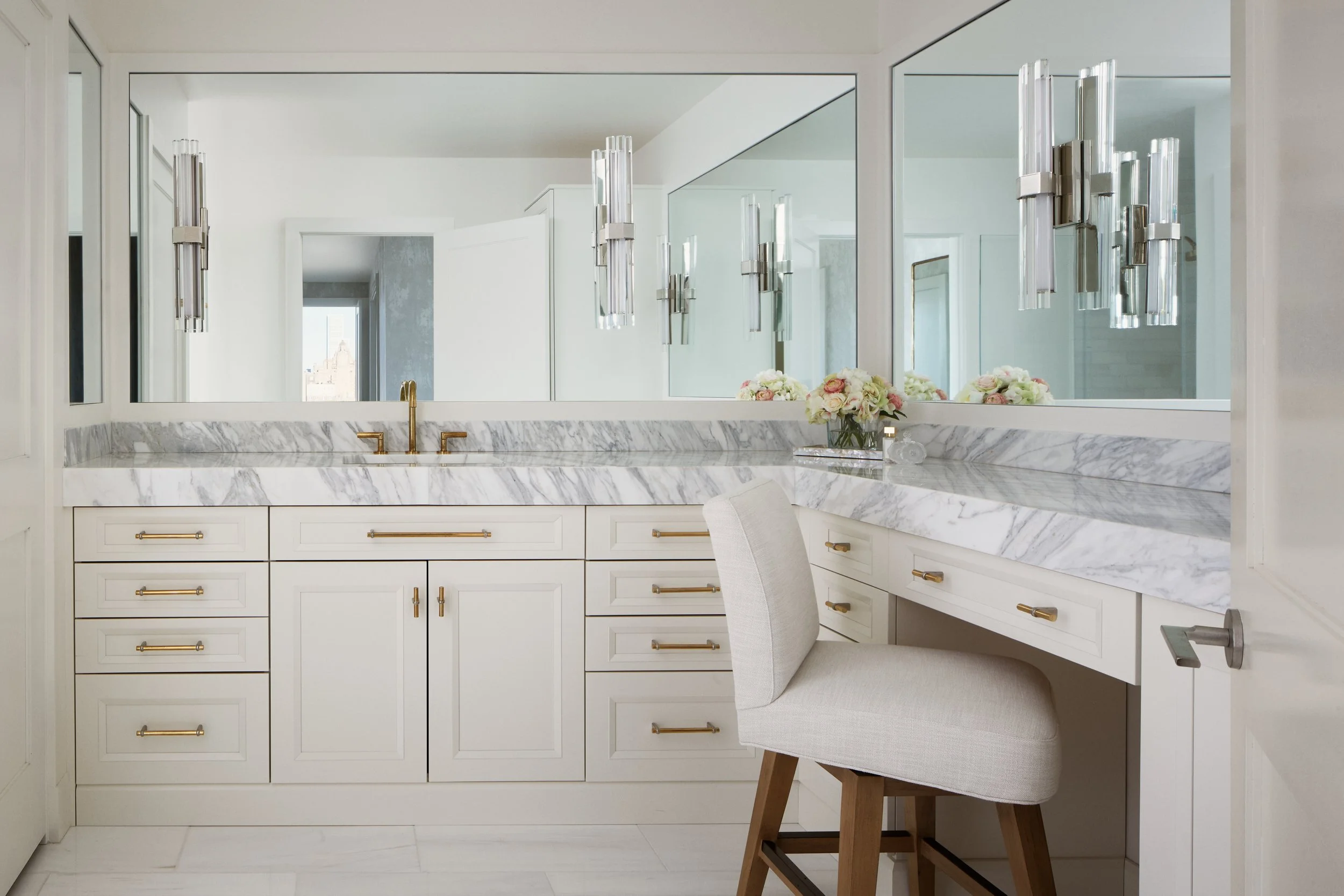 A luxurious bathroom vanity area with a large mirror, white cabinetry with gold handles, a marble countertop, and modern wall sconces. There is a chair and a bouquet of flowers on the counter.