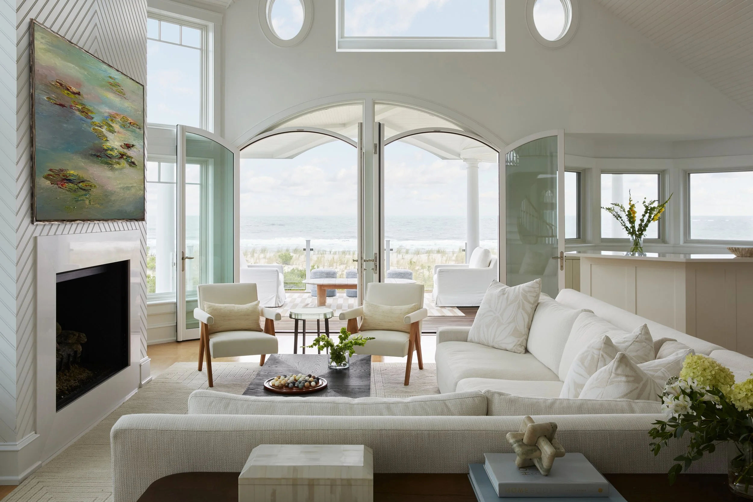 Living room with white sofa, two chairs, fireplace, and large windows with ocean view, open doors leading to a porch with outdoor seating and ocean scenery.