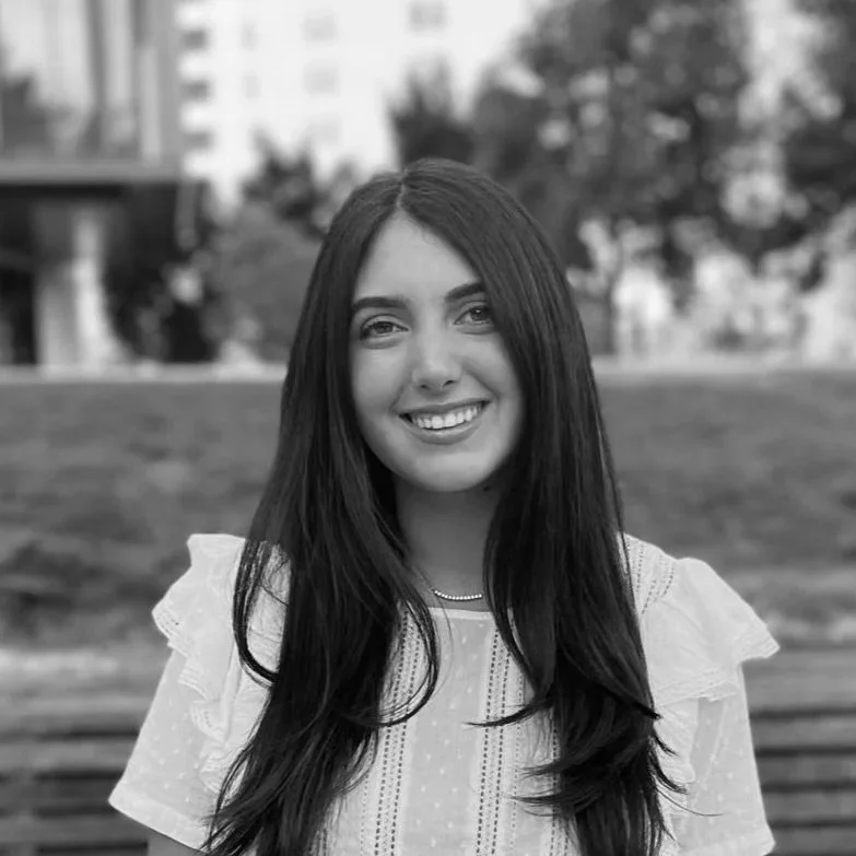 A young woman with long dark hair smiling outdoors in a park with buildings and trees in the background.