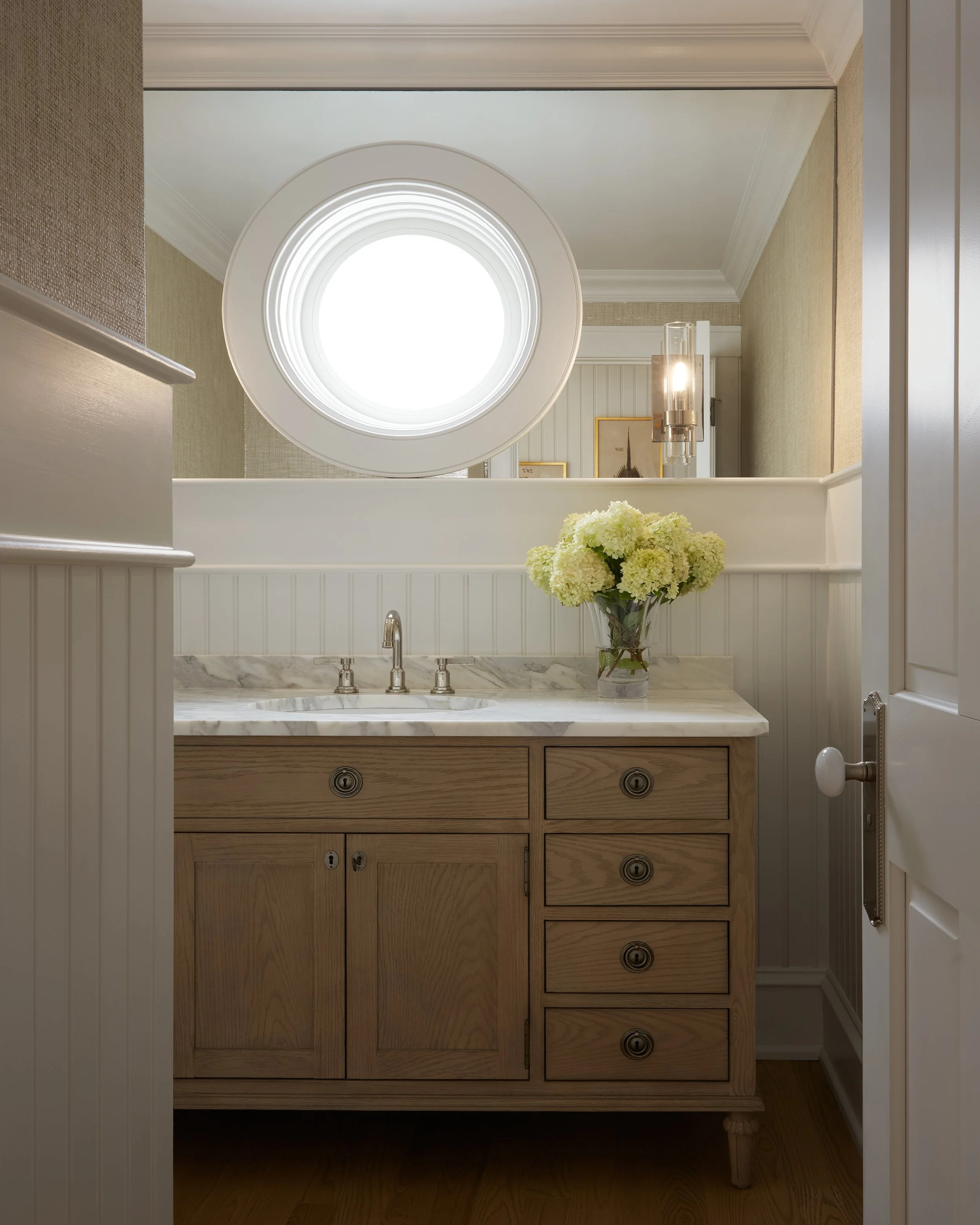 Bathroom vanity with a wooden cabinet, marble countertop, and a bouquet of white flowers, with a round mirror and window above.