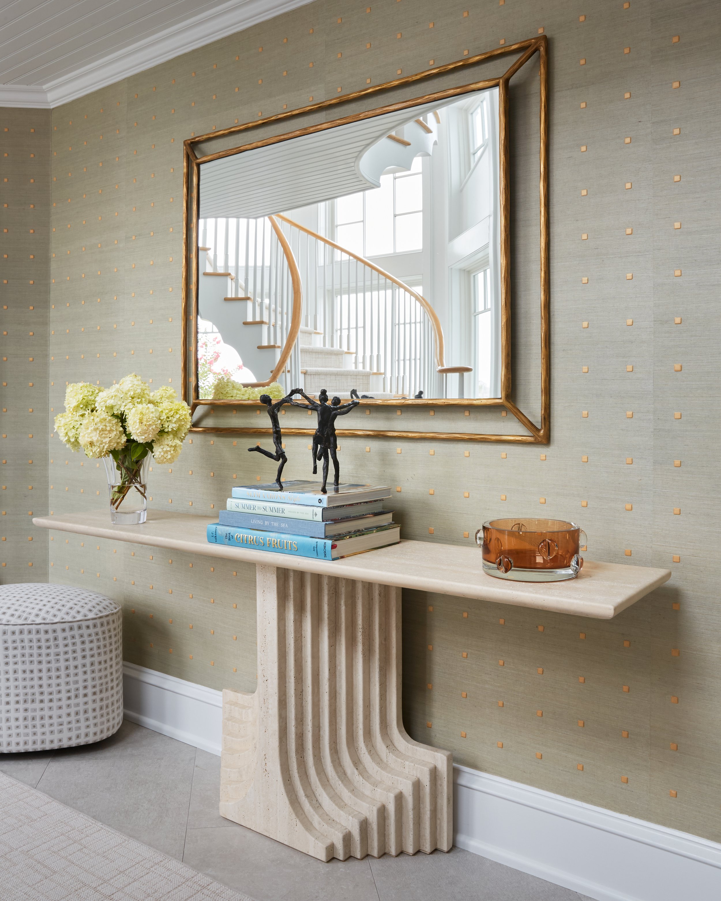 A beige console table with a fluted base, decorated with a glass vase of white hydrangeas, a stack of books, a small black sculpture, and an amber glass bowl, all reflected in a large gold-framed mirror on a patterned wall.