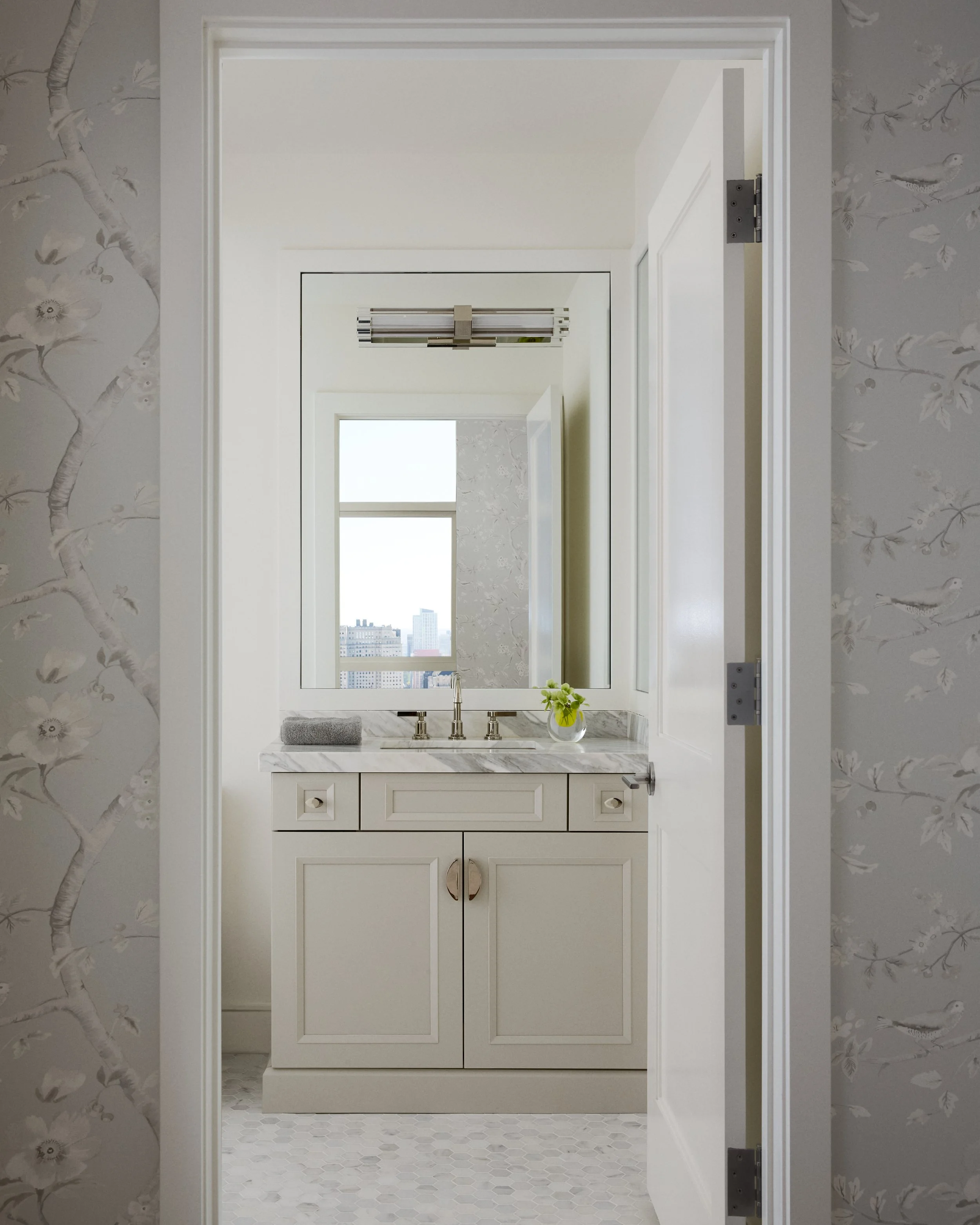 Bathroom vanity with white cabinet, marble countertop, silver faucet, small plant, mirror, window in the background, and floral wallpaper on the walls.