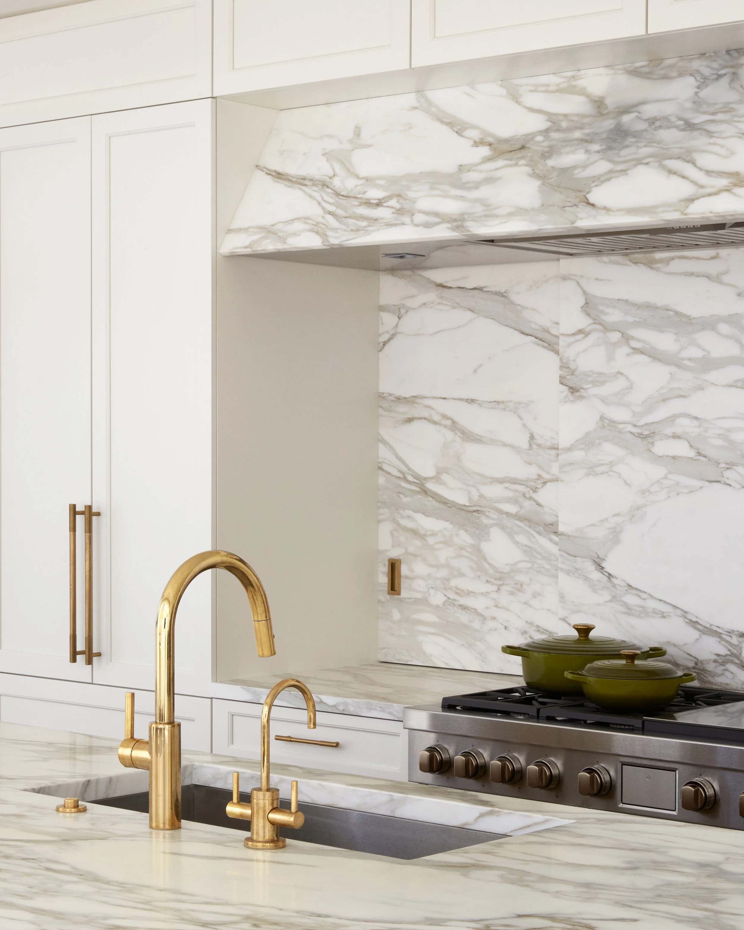 Modern kitchen with white marble countertops and backsplash, gold faucet, and green pots on stove.
