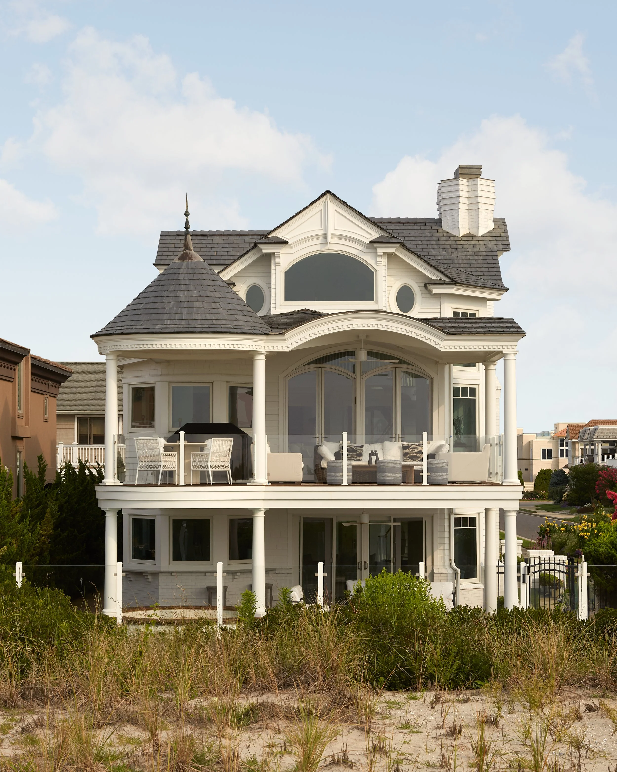 A large, Victorian-style beach house with white exterior, multiple porches, and a turret, situated near the beach with grass in the foreground.