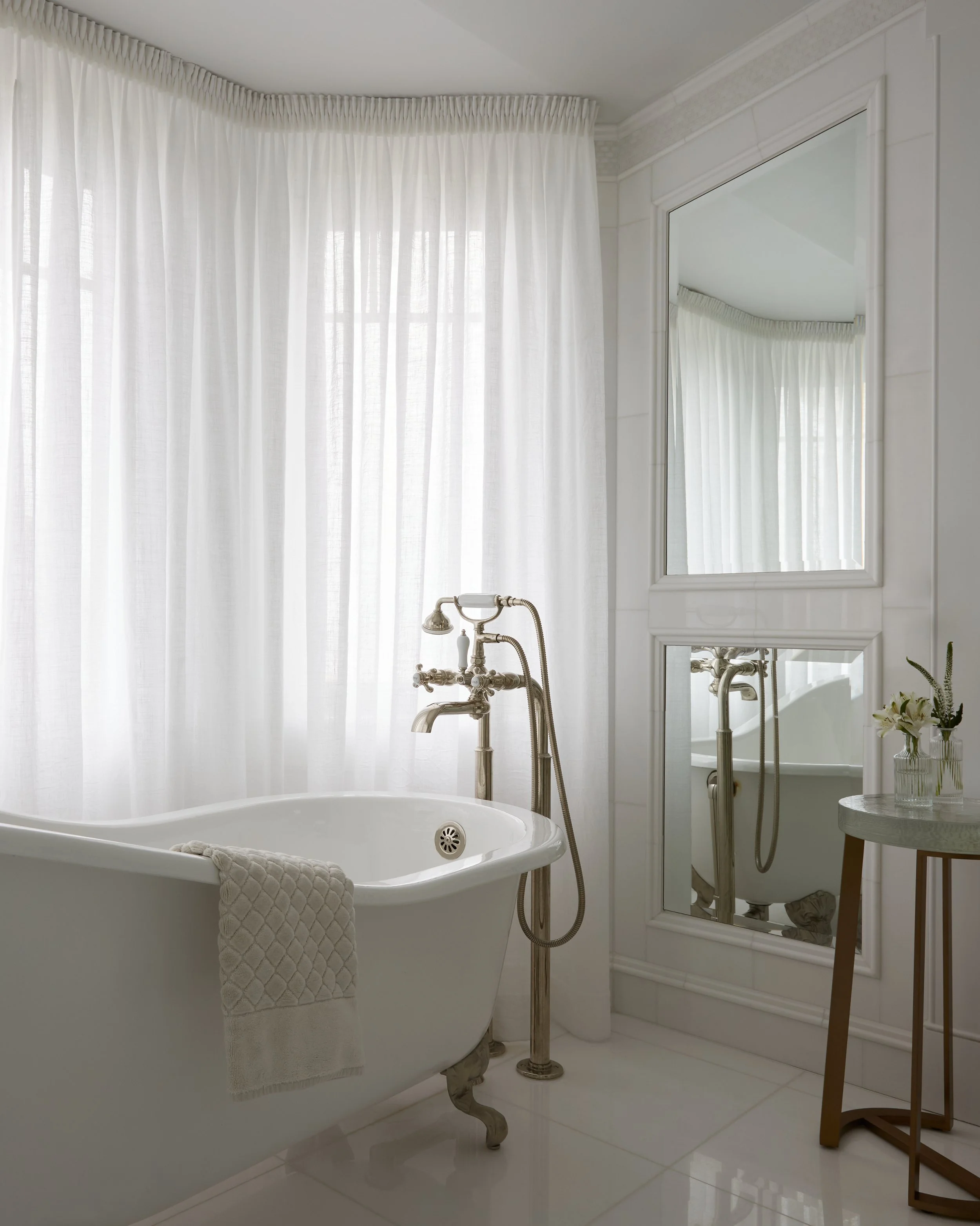 A bathroom with a vintage bathtub, surrounded by white curtains and walls, with a large mirror reflecting the bathroom fixtures and a small table holding a vase with flowers.