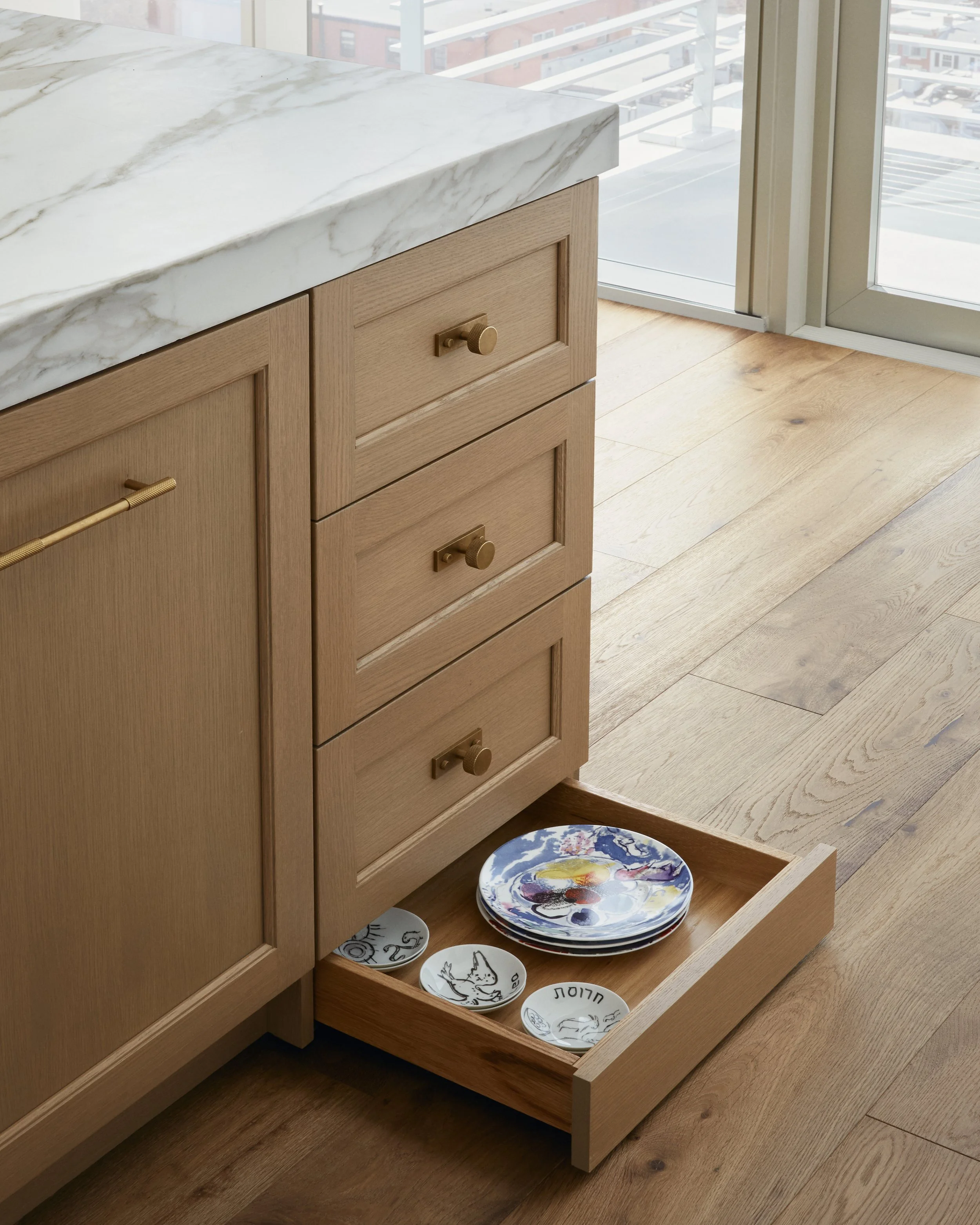 Wooden kitchen cabinet with a marble countertop, open drawer with decorative plates on a wooden floor, next to sliding glass door.