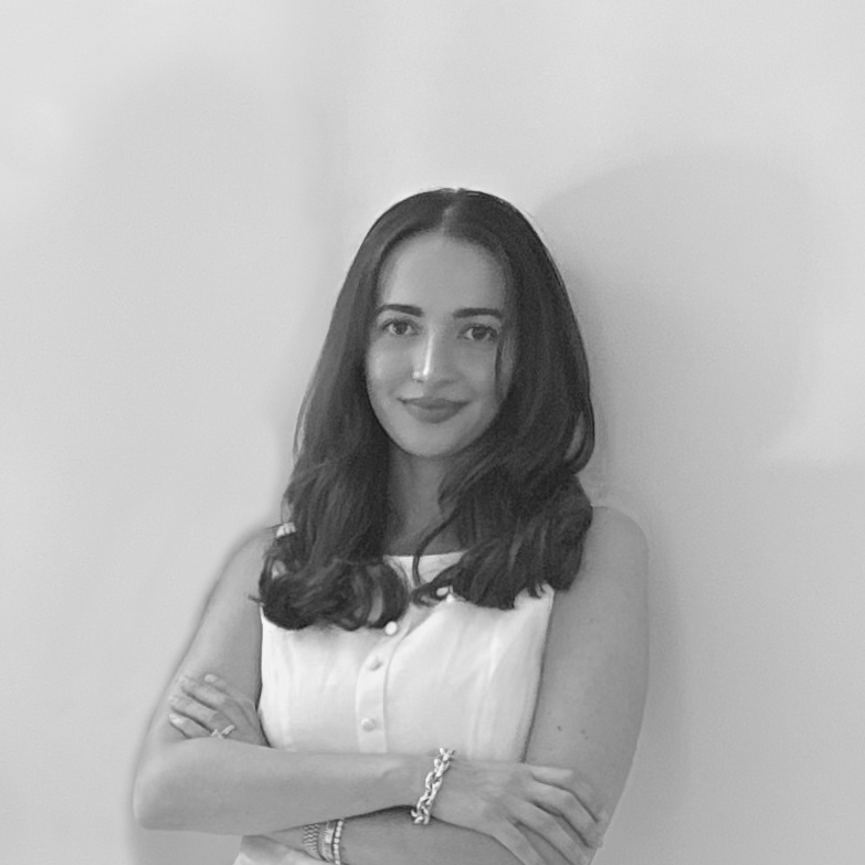 Black and white photo of a woman with dark, wavy hair, wearing a sleeveless button-up top, standing with her arms crossed against a plain wall.