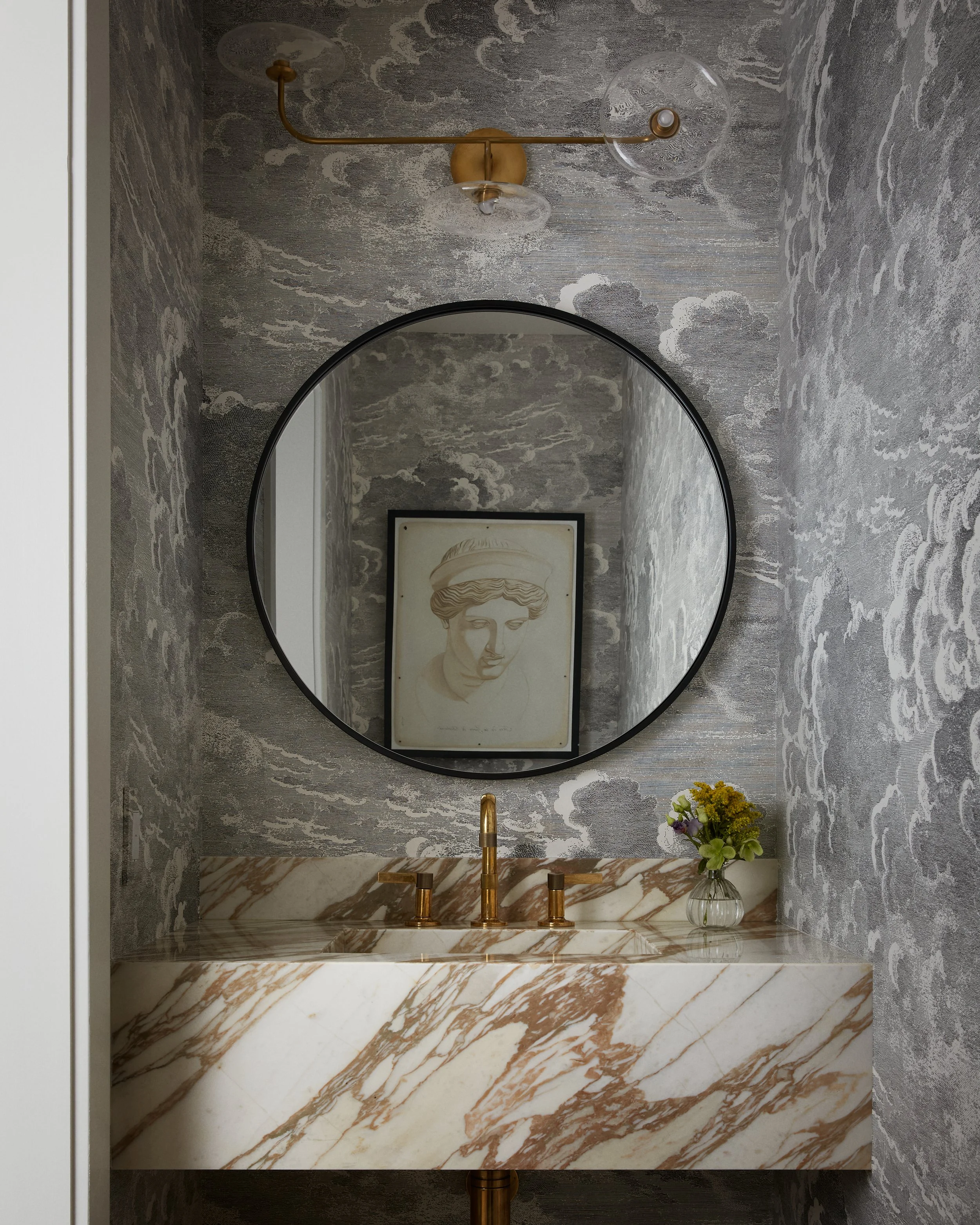 Bathroom vanity with marble countertop, gold faucet, round mirror, textured gray walls, artwork, and a small vase of flowers.