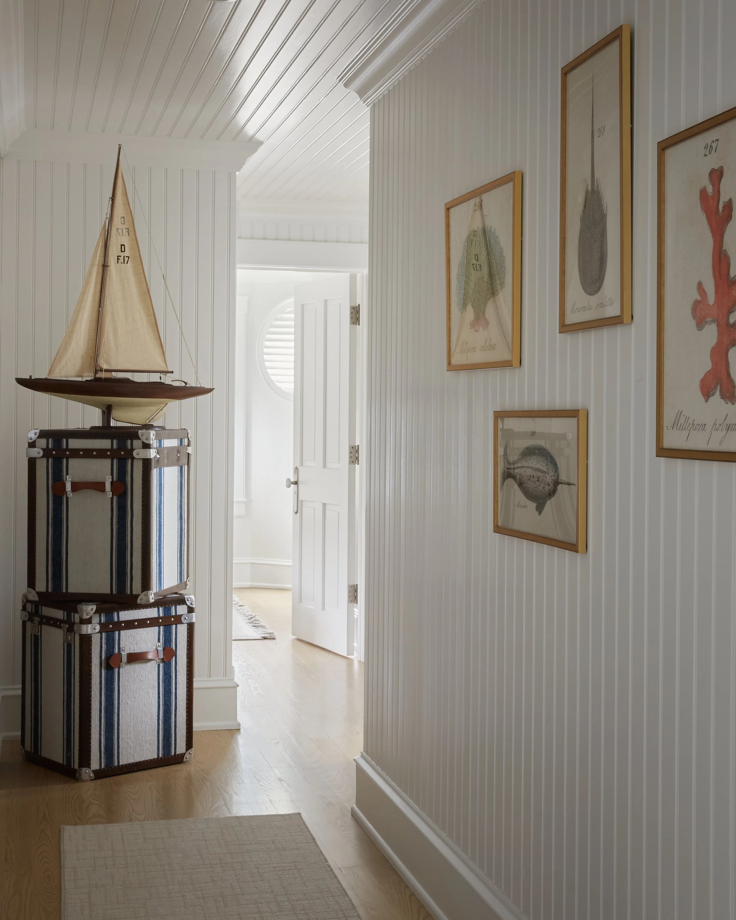 A white hallway with wall art and a model sailboat on top of striped storage trunks near an open door with white shutters.